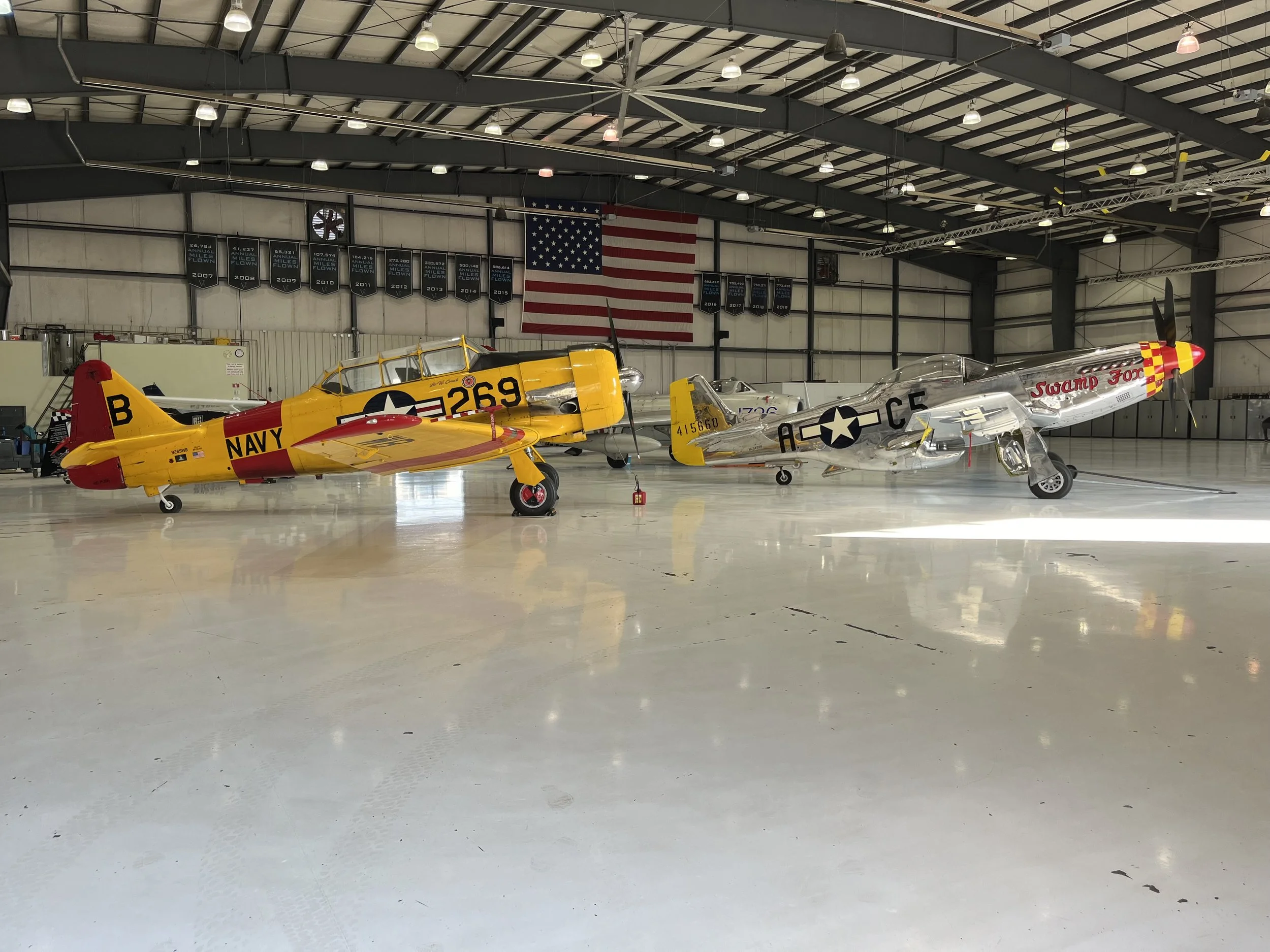 Three vintage aircraft inside a hangar with an American flag on the wall.