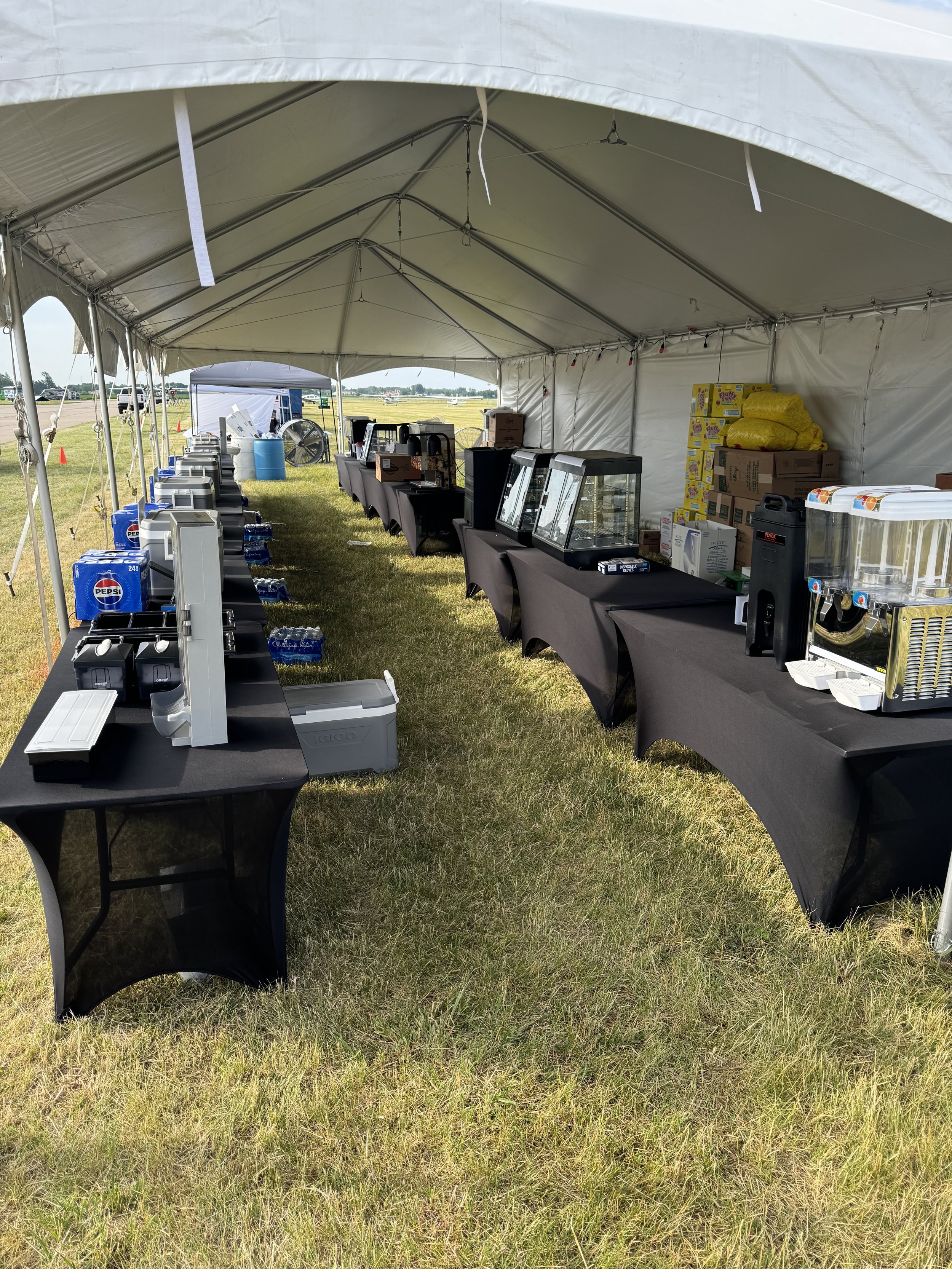 Food and drink station under a large event tent, with tables covered in black tablecloths displaying drink dispensers, snack boxes, and emergency supplies, set up on grassy ground.