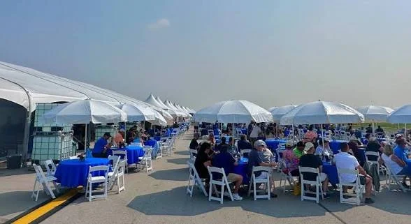 Outdoor event with guests sitting under white tents and umbrellas, blue tablecloths, and white chairs