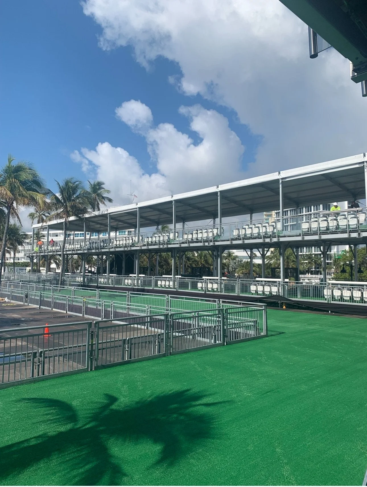Empty grandstand and green court at a tennis stadium with palm trees and a partly cloudy sky in the background.