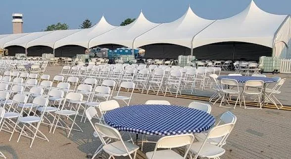 Outdoor event setup with round tables covered in blue and white checkered tablecloths, surrounded by white folding chairs, under large white tents.