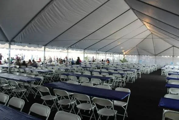 Empty large event tent with rows of folded chairs and tables, some with dark tablecloths, set up for a gathering or celebration, with people visible inside.