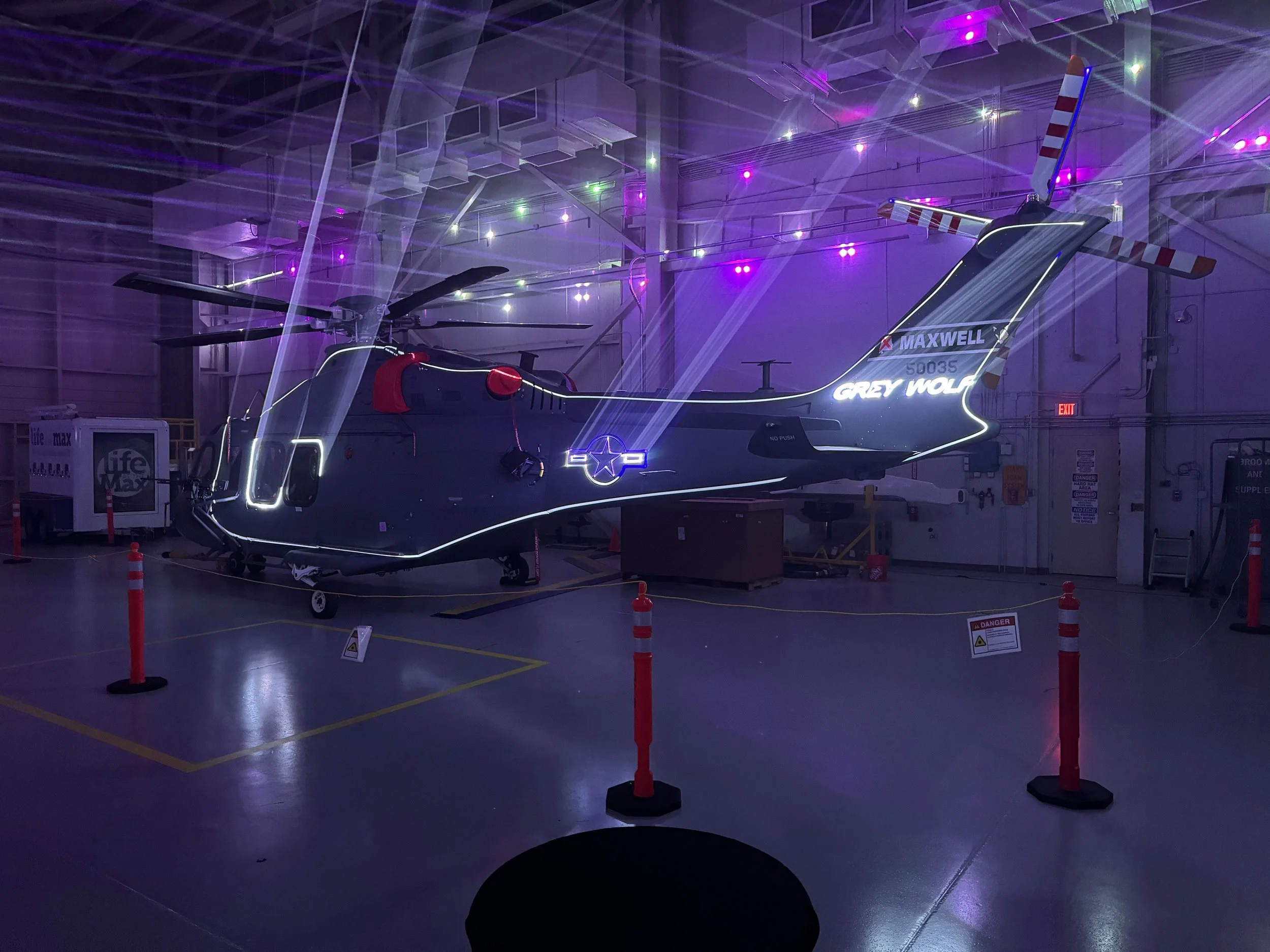 A helicopter with neon lights inside a hangar, illuminated with purple and white lights, surrounded by safety cones and warning signs