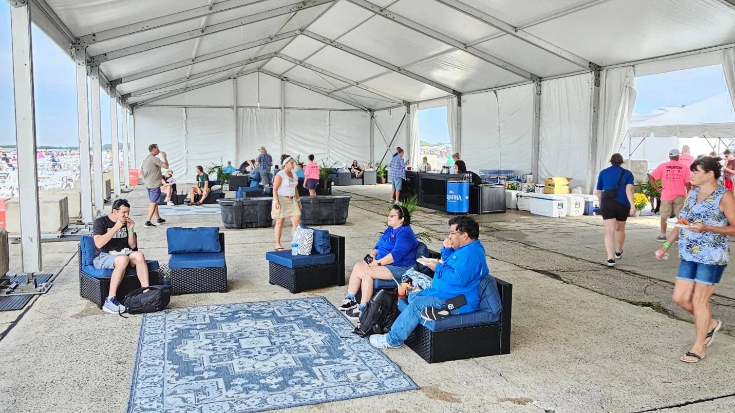 People sitting and walking inside a large white tent at a daytime outdoor event, with beach and ocean visible outside the tent.