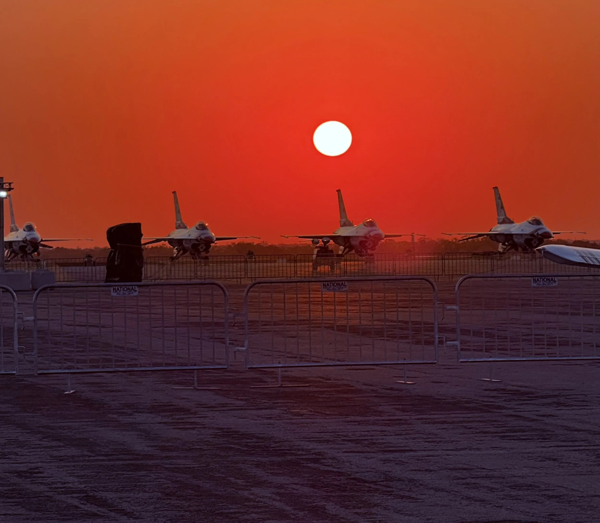 Four fighter jets parked on a runway with a fence in front, backdrop of a bright orange sunset.