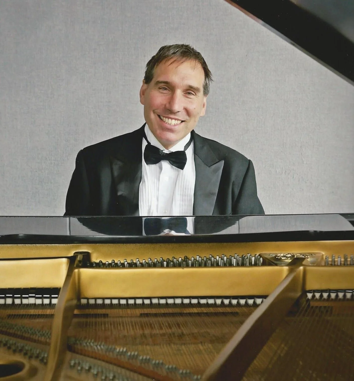 A man in a tuxedo sitting at a grand piano, smiling, with the piano strings visible in the foreground.