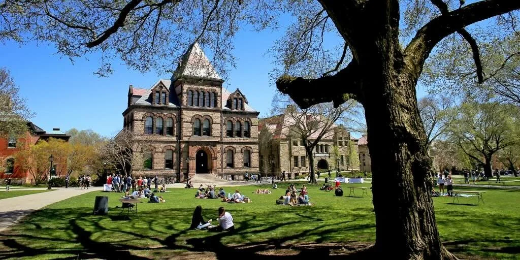 Brown University campus with historic stone building, students sitting on the lawn, and large tree in the foreground