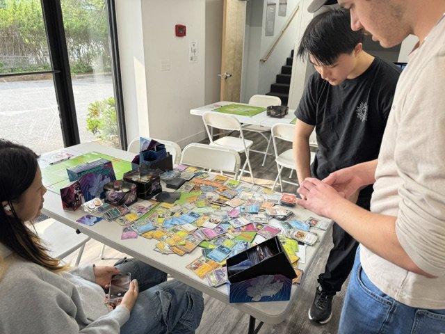 Three people playing a card game with colorful cards spread across a white table in a bright room with large windows.