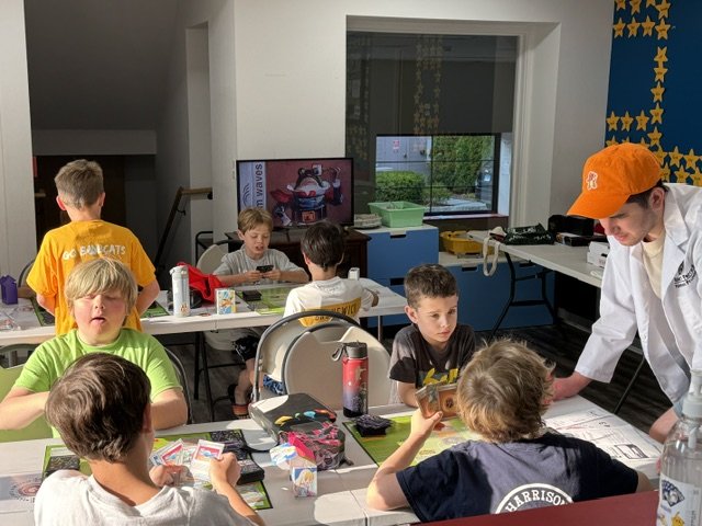 Kids playing card games at a table in a classroom or indoor activity room with an adult assisting.