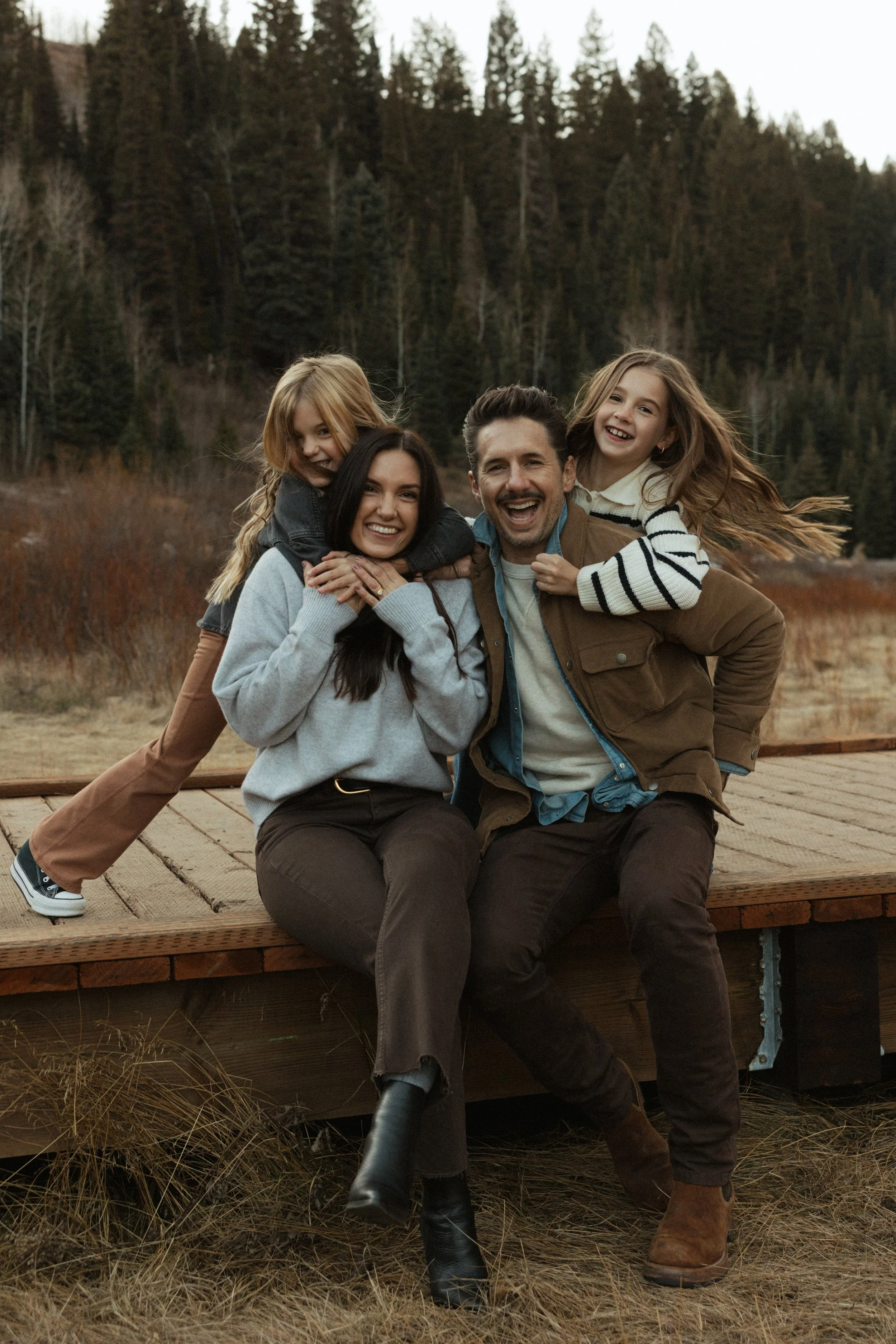 A happy family of four outdoors in a wooded area, with two children riding on their parents' backs and smiling, during daytime.