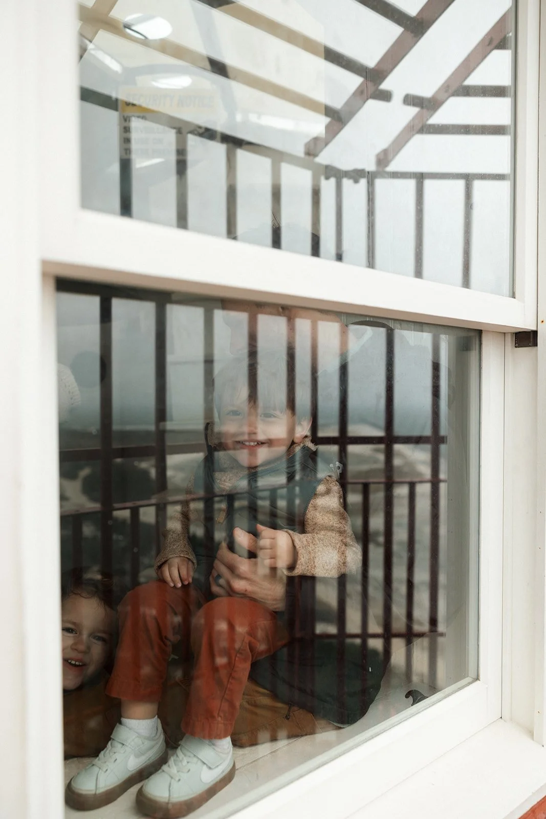 Two children sitting inside behind a glass window, with one smiling and holding a device, seen through reflections on the glass.