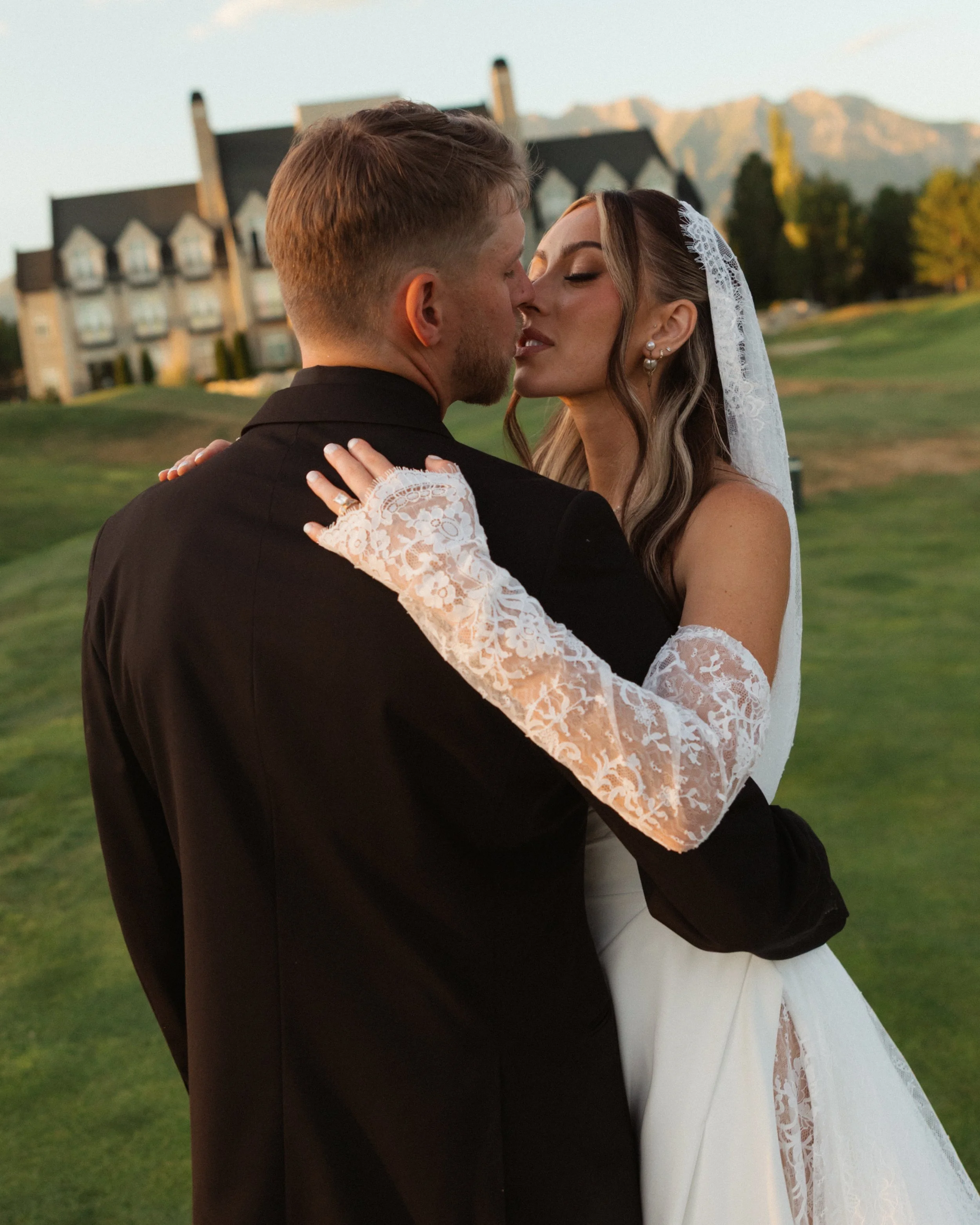 A bride and groom kissing outdoors on a grassy field, with a large building and mountains in the background during sunset.