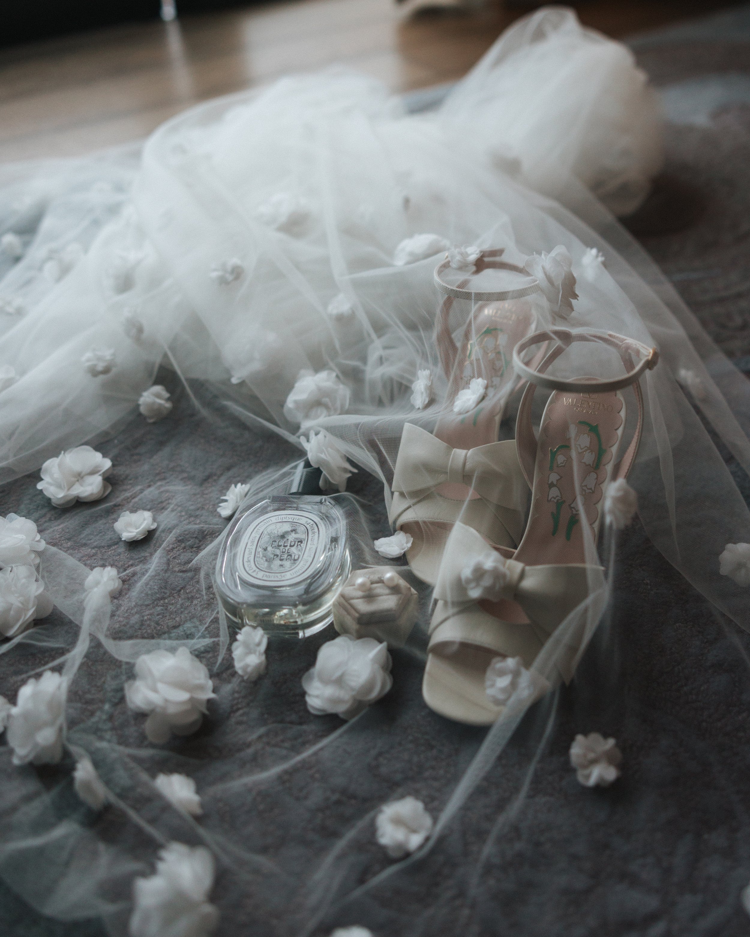 Wedding accessories including a white wedding dress, a pair of high-heeled shoes with bows, a small perfume bottle, a jewelry box, and scattered flower petals and ribbons on a dark fabric surface.
