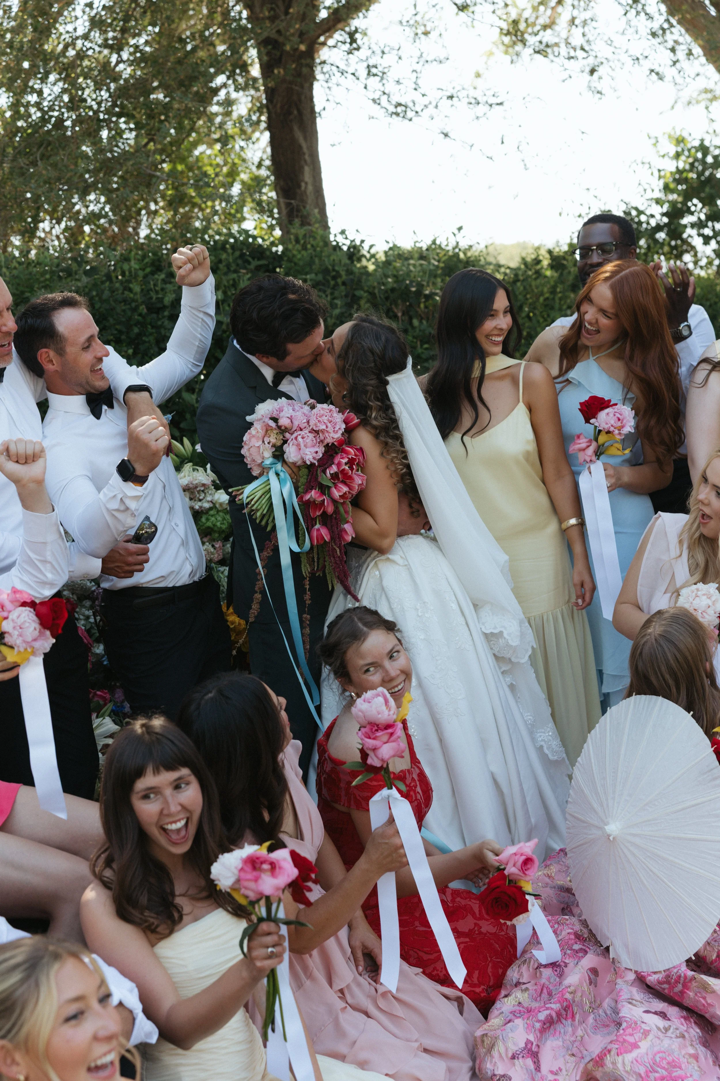 A group of people celebrating at a wedding, with the bride and groom kissing and friends holding flowers and white parasols.