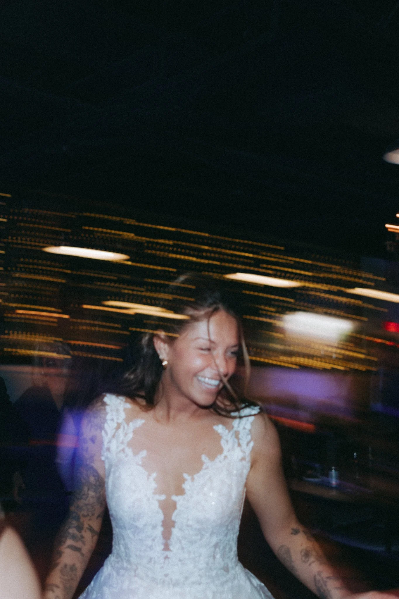 A woman in a white lace wedding dress is smiling and winking at a celebration or party, with blurred colorful lights in the background.