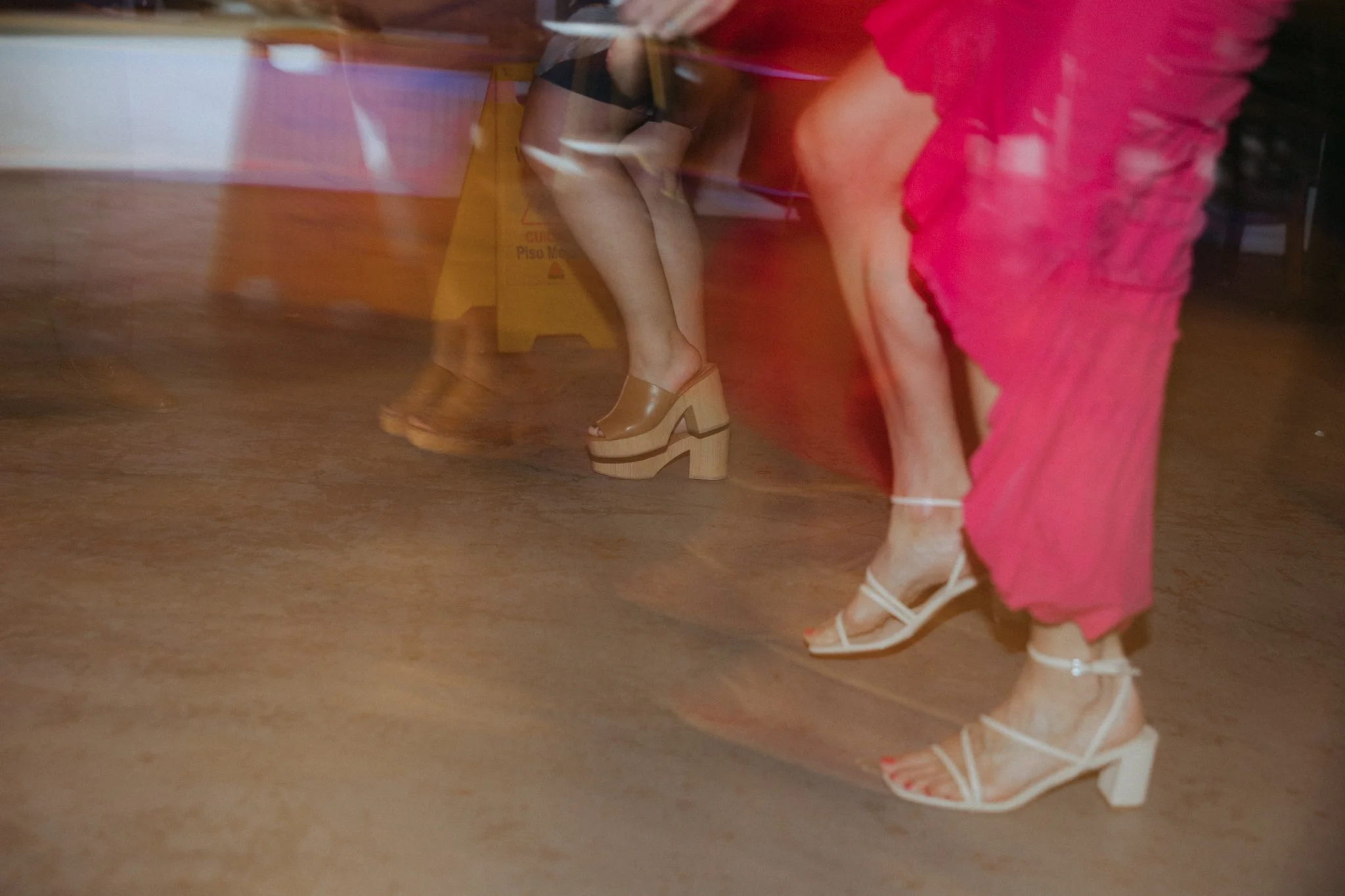 People sitting at a table with their legs and shoes visible, captured with motion blur, including a woman in a pink dress and white high-heeled sandals.