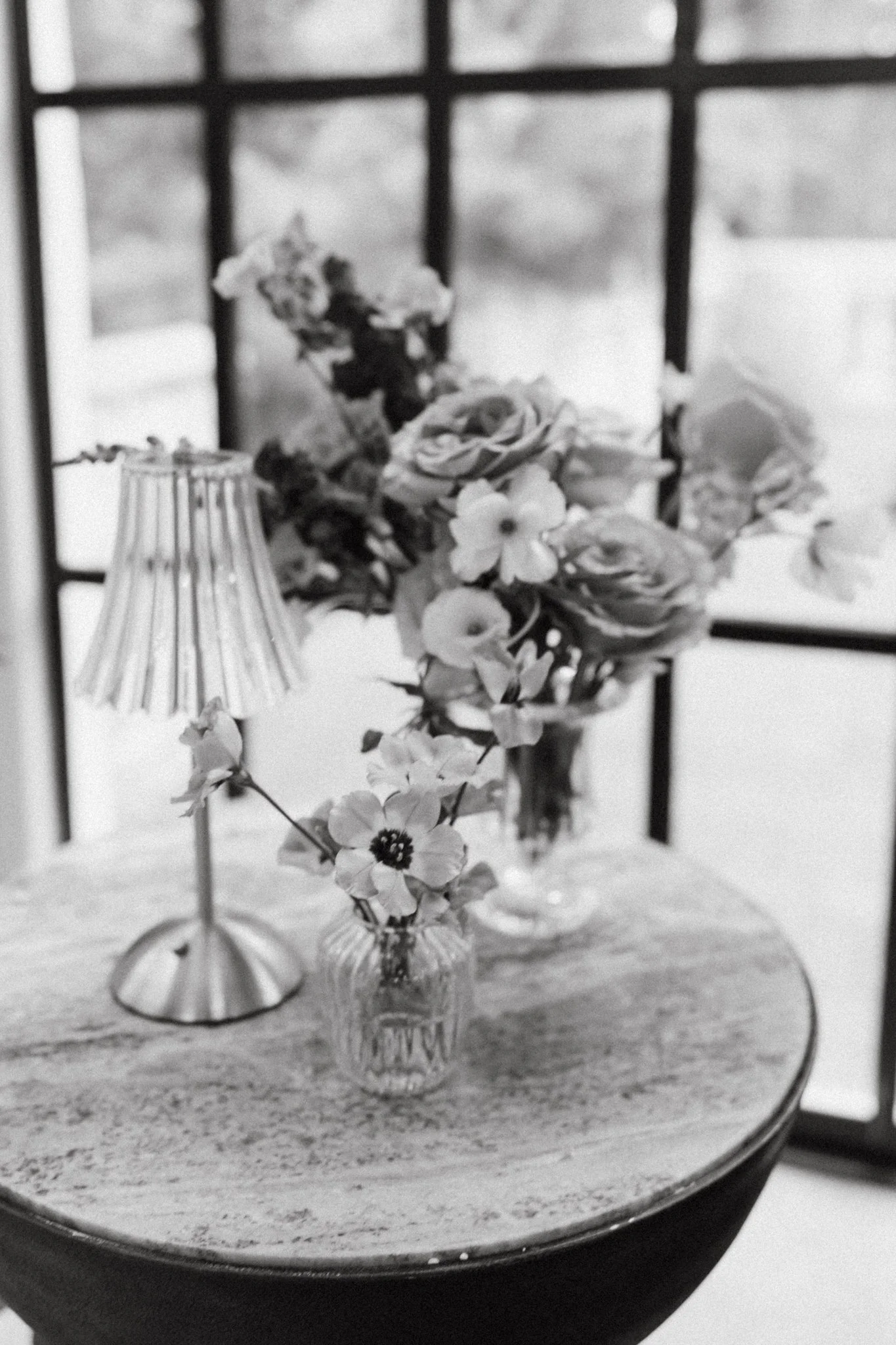 A black and white photo of a small round table with a floral arrangement in a jar, a lamp, and a window with grid panels in the background.
