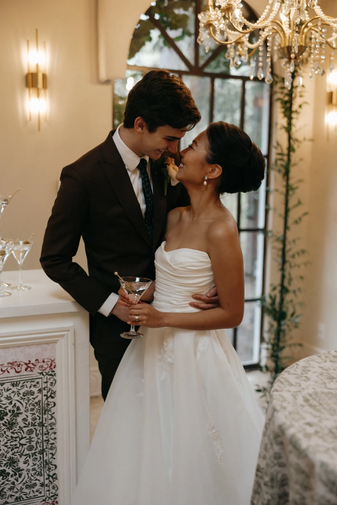 A bride and groom sharing a moment at their wedding reception, smiling and looking into each other's eyes, with the bride holding a cocktail glass.