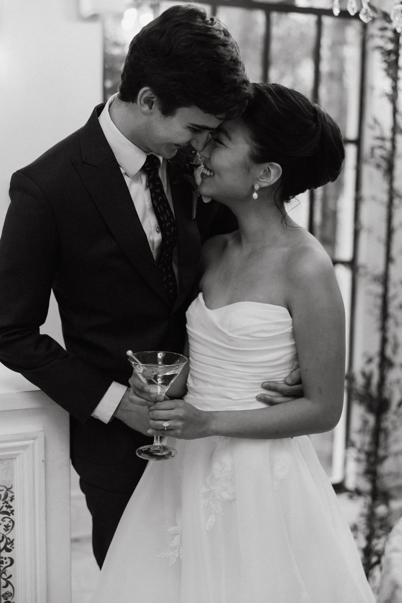 A black and white photo of a couple at a wedding, standing close, smiling and touching foreheads. The woman is in a strapless wedding dress holding a martini glass, and the man is in a suit and tie, leaning towards her.