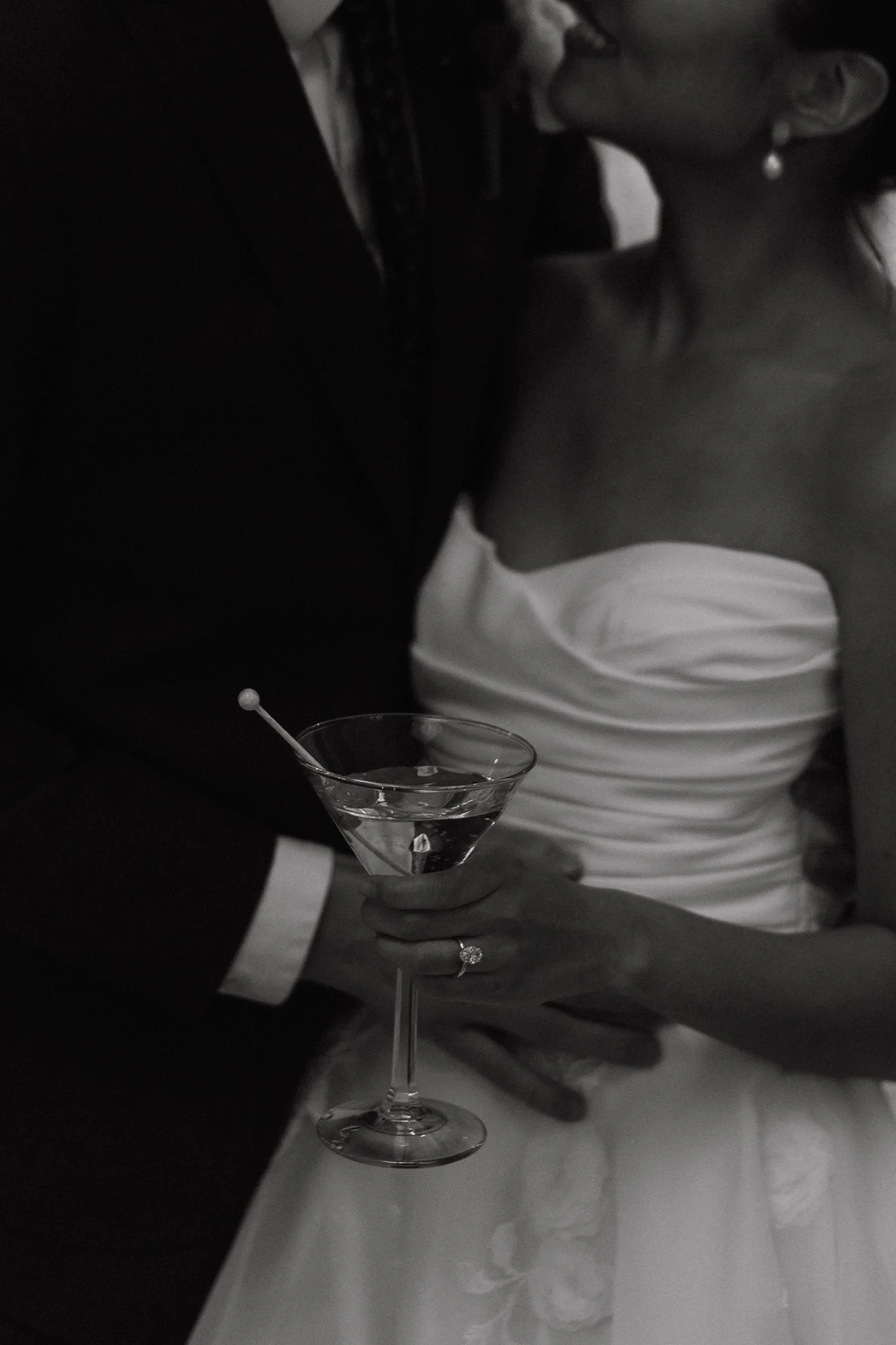 Close-up of a bride and groom at a wedding, with the groom holding a cocktail glass in front of the bride, who is wearing a strapless wedding dress, in a black-and-white photo.