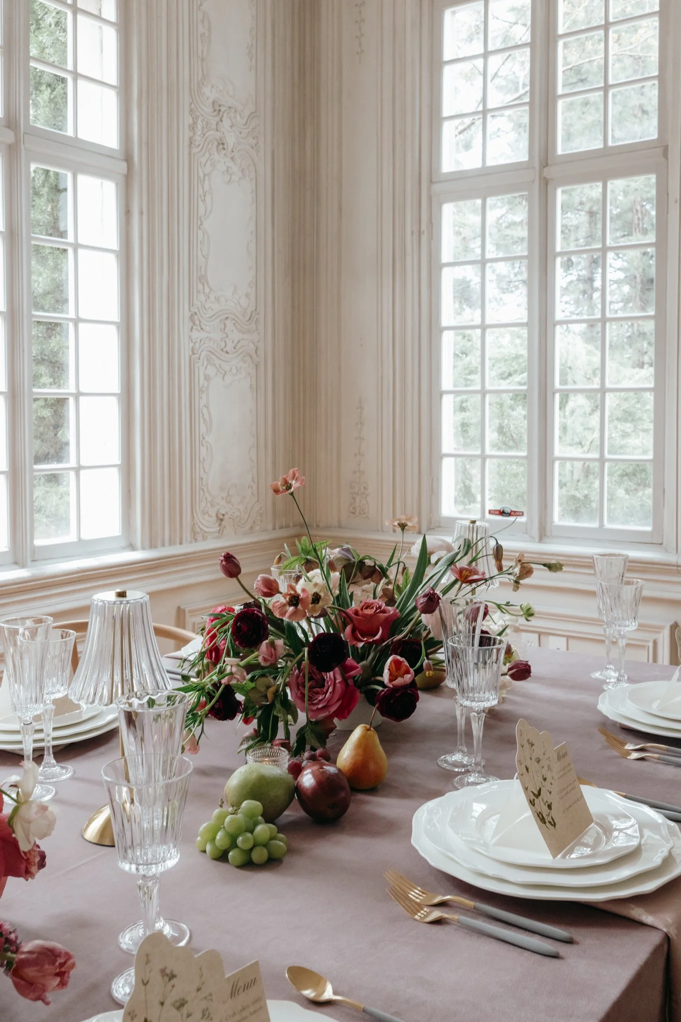 Elegant dining table set with white plates, gold utensils, crystal glasses, a flower arrangement with pink, red, and black flowers, and assorted fruit including pears, grapes, and apples, in a bright room with large windows.