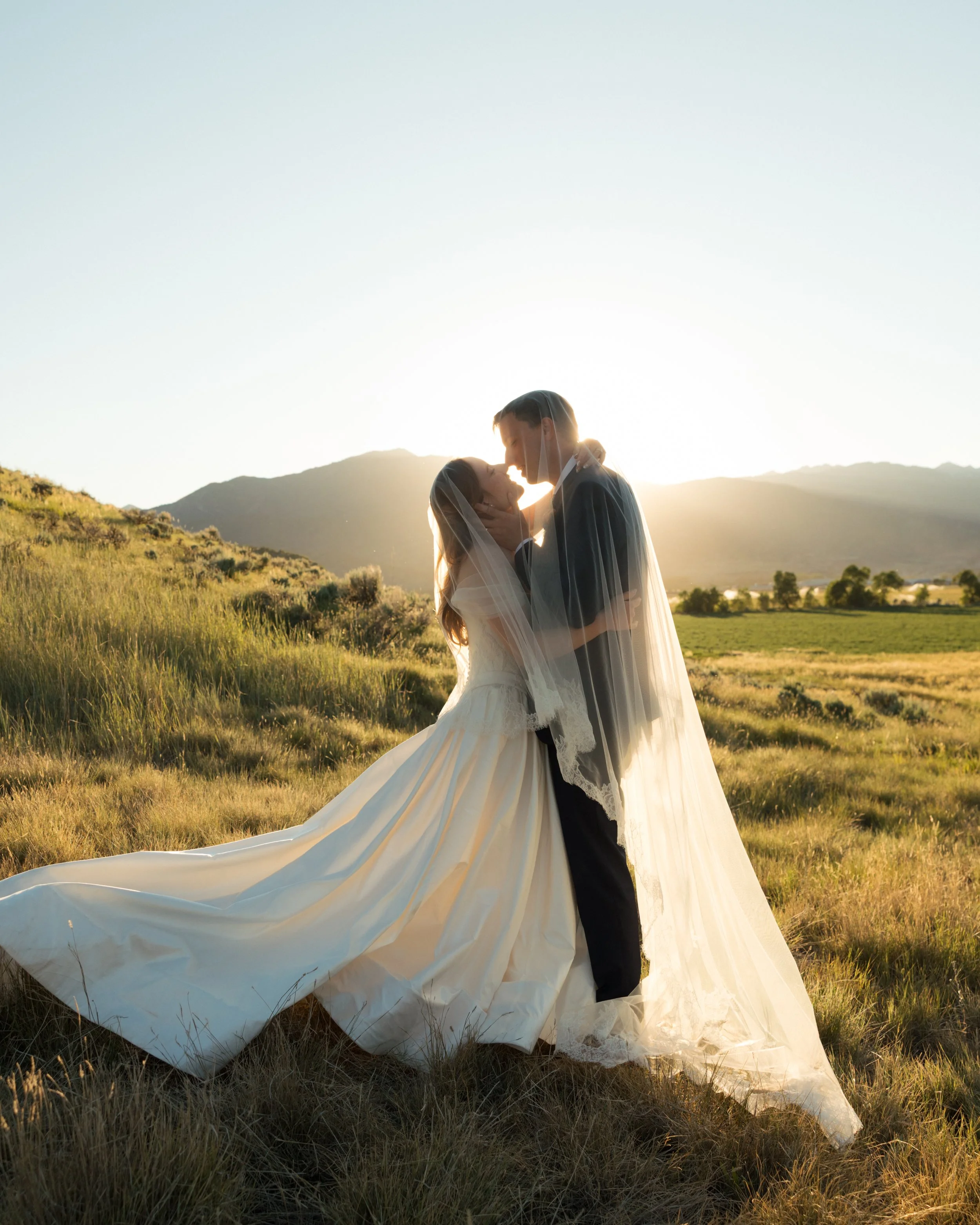 A bride and groom embracing in a field at sunset, with the bride's white wedding gown flowing on the ground and the groom dressed in a suit, mountains in the background.