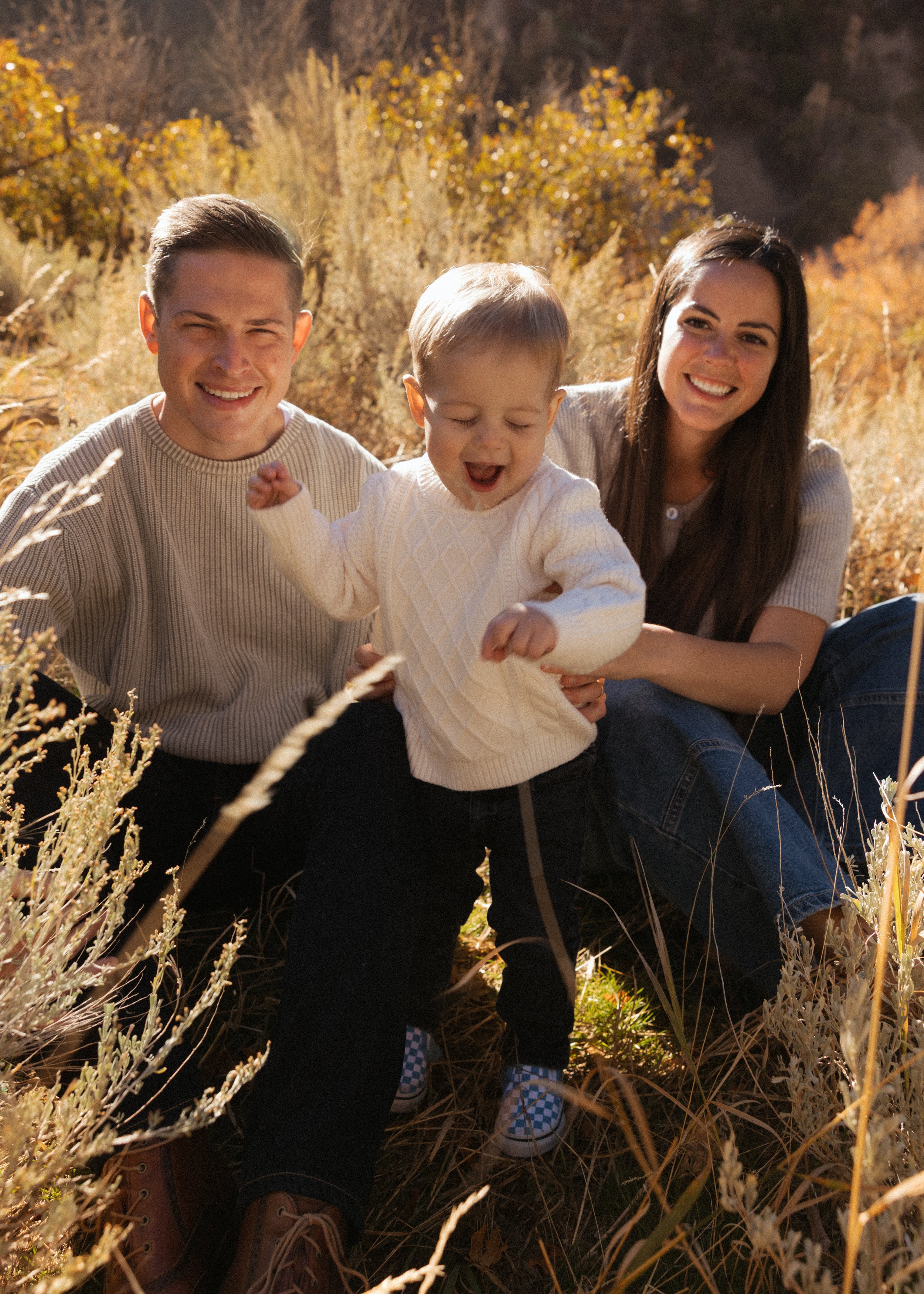 A happy family of three, a man, a woman, and a young child, sitting outdoors in a grassy field during fall. They are smiling and enjoying the sunny weather with autumn-colored trees in the background.