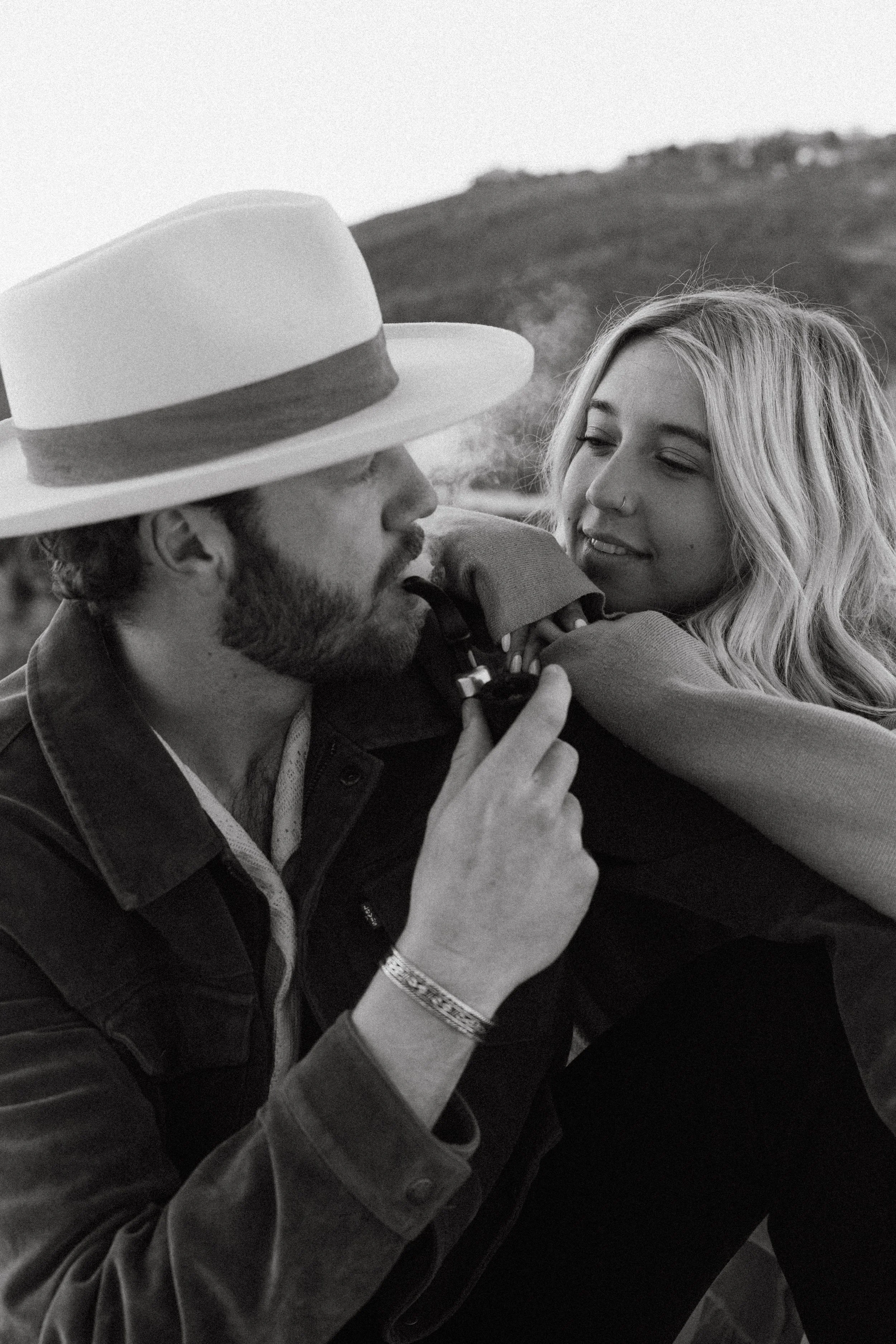 A black and white photo of a man with a beard and a woman with long hair, both looking at each other and smiling, with a blurred natural background.