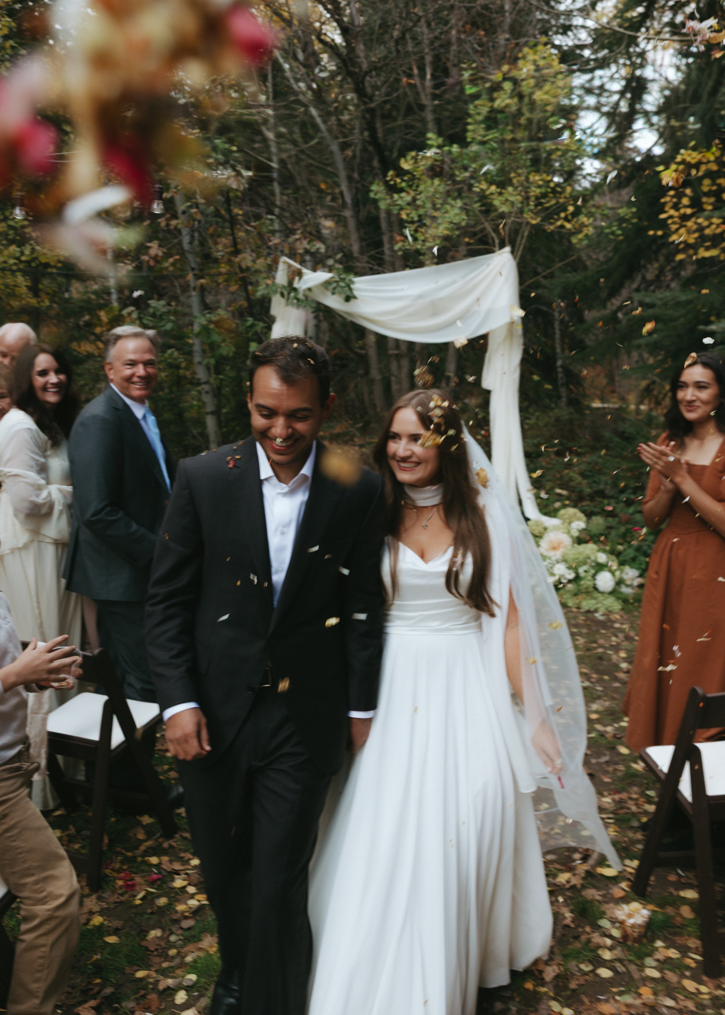 A newly married couple walking outdoors under a wedding arch, surrounded by smiling guests, with confetti falling around them, in a forest setting with trees and autumn leaves.