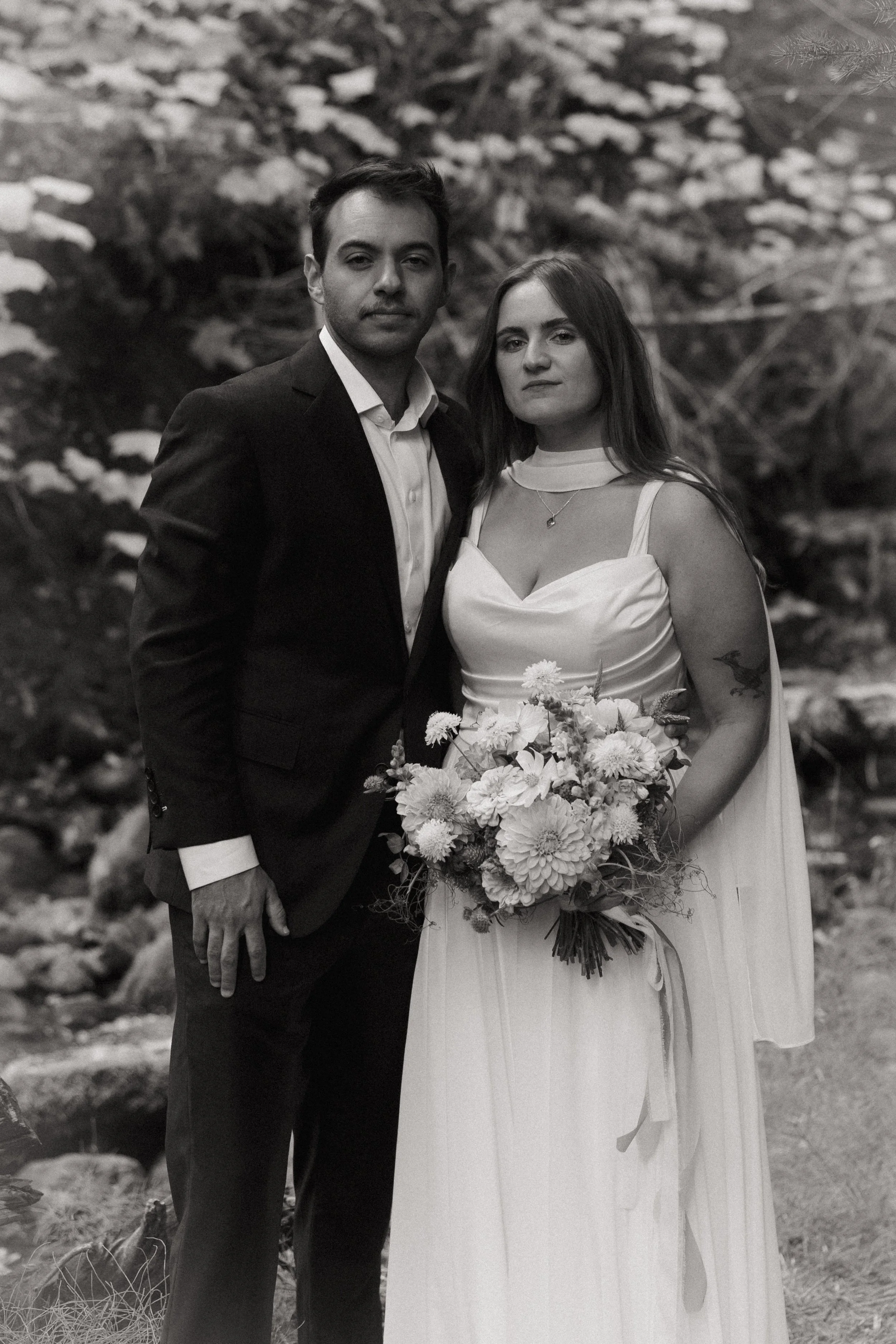 Black and white photograph of a man and woman at a formal outdoor event, the woman holding a bouquet of flowers, both looking at the camera with serious expressions.