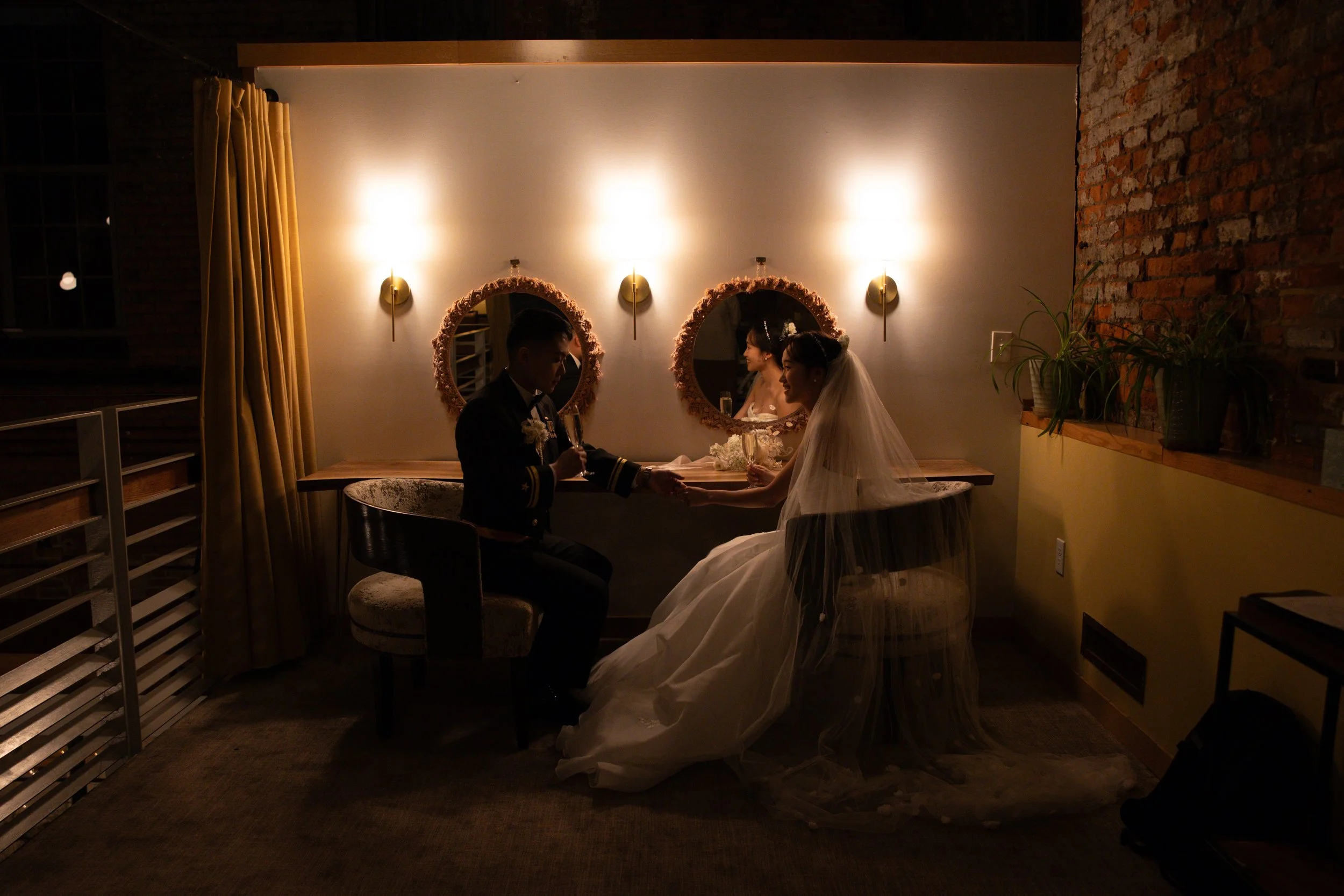 A bride and groom holding hands and facing each other at a dimly lit indoor wedding reception, with two mirrors behind them and three wall-mounted lamps above.