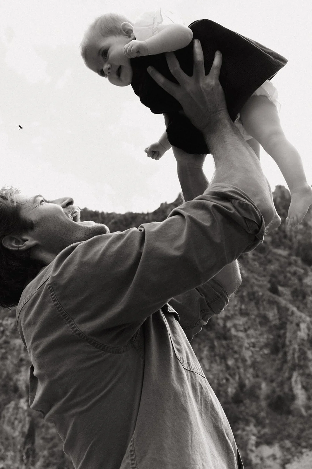 A man lifts a young girl into the air, both smiling and looking at each other outdoors with trees in the background.