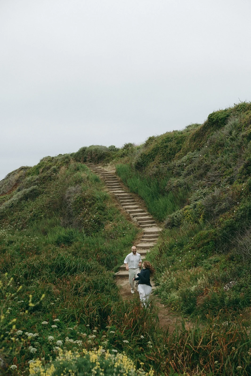 Two people walking on a trail on a hillside with outdoor stairs, surrounded by green plants and bushes, under an overcast sky.