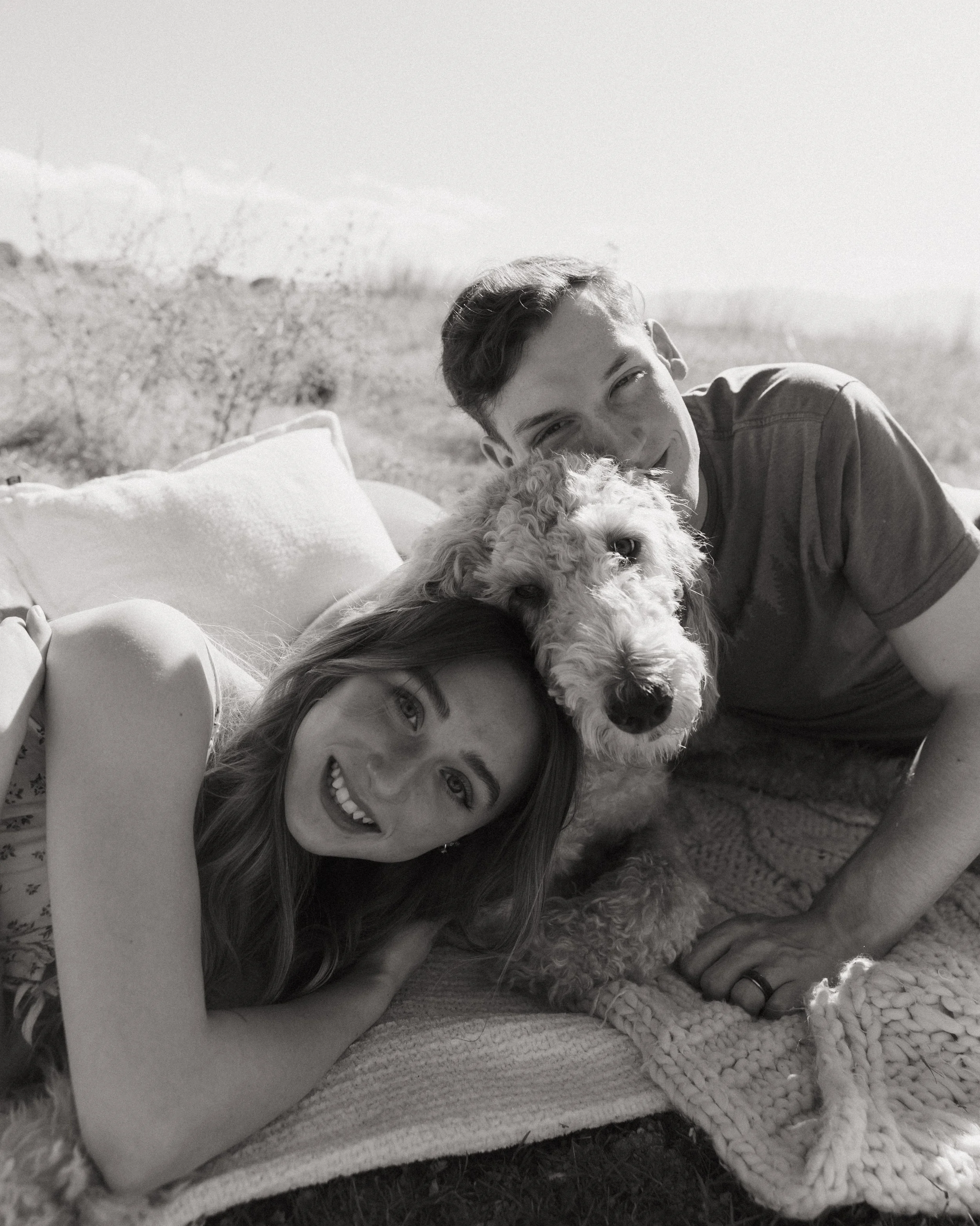 A young woman is lying on a blanket outdoors, smiling at the camera, with a boy and a fluffy dog nearby. The boy is partially visible, resting his head on the blanket, and the dog is lying close to the girl, looking up with a relaxed expression.