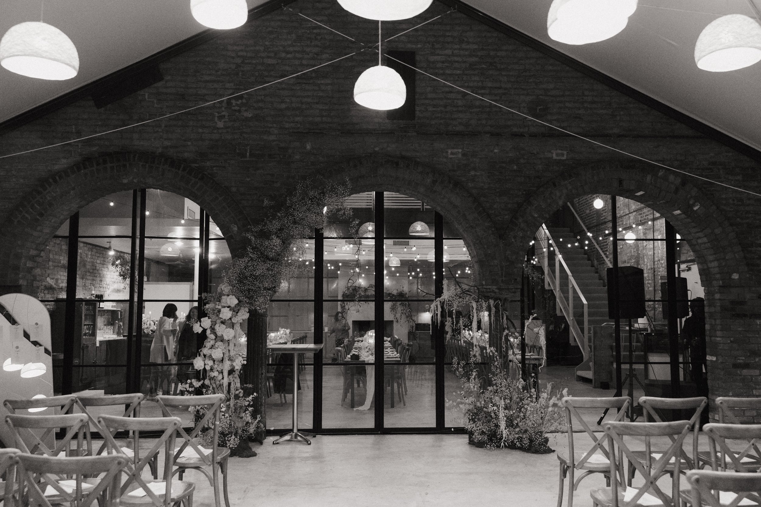 Interior of a decorated event hall with floral arrangements, string lights, and a long table set for a gathering, viewed through large glass doors, with chairs arranged in front.