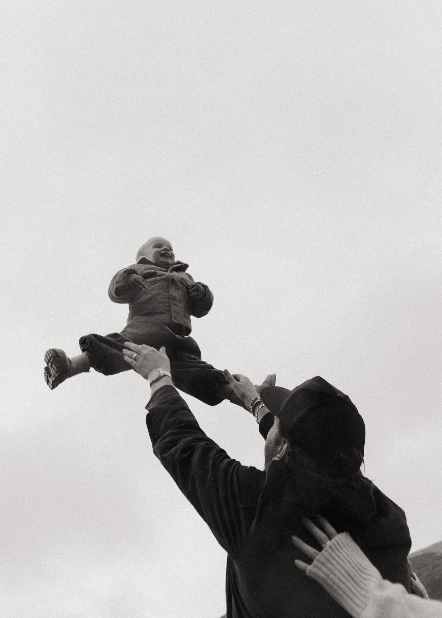 A person wearing a cap lifting a smiling child into the air against a plain sky