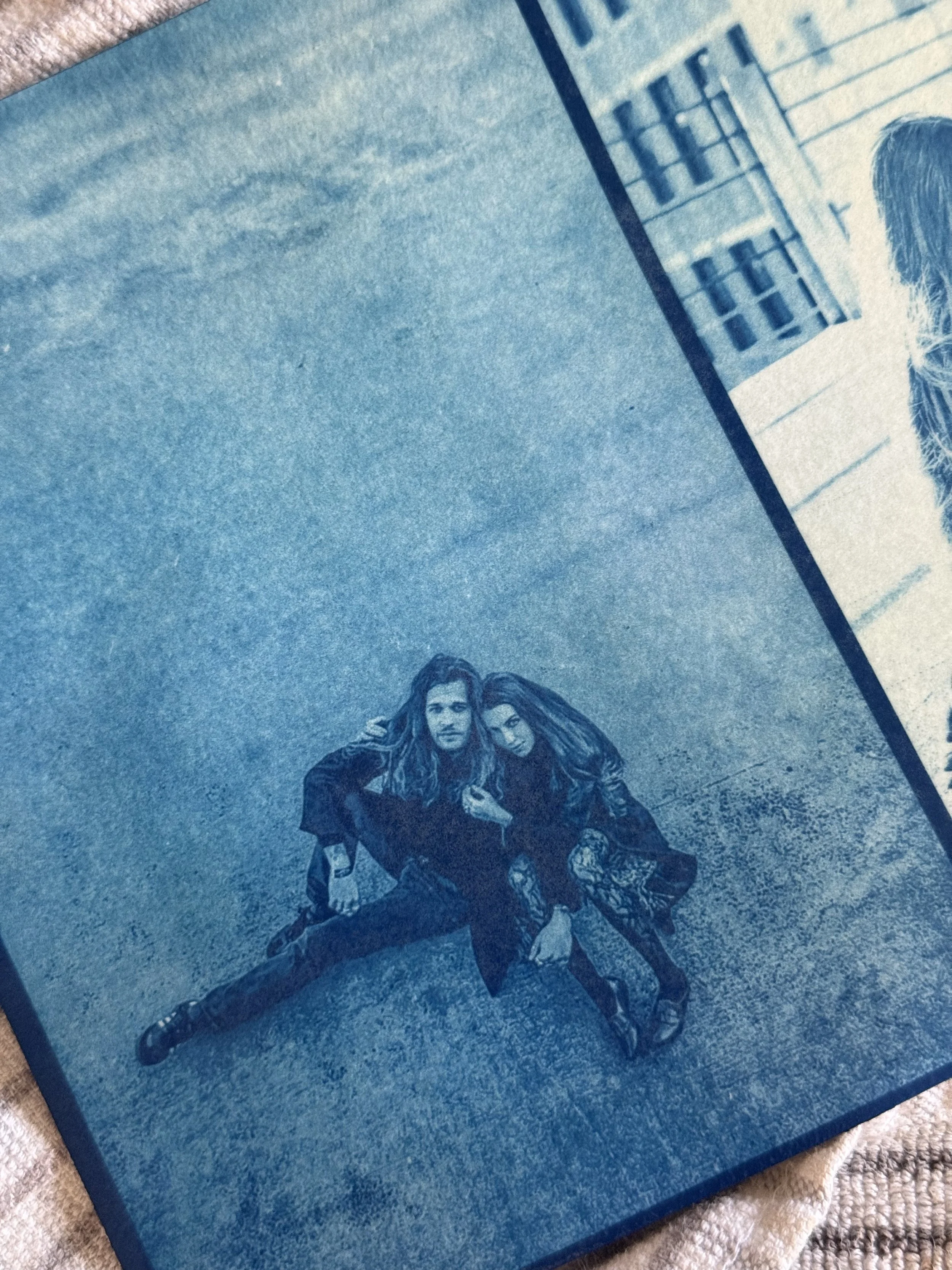 A blue-toned image on a printed surface featuring two young people sitting on the ground, a man with long hair and a woman with long hair, both dressed in dark clothing.
