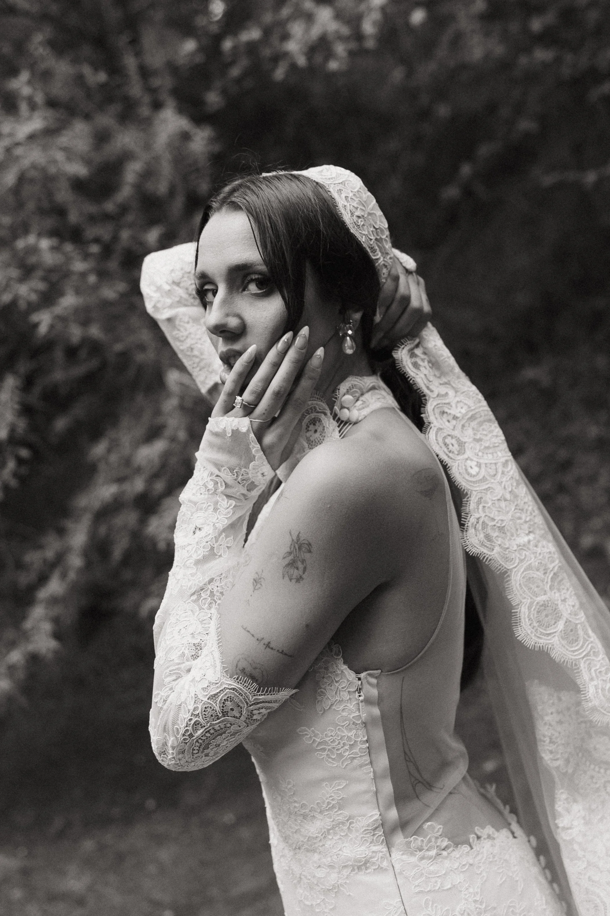 Black and white photograph of a woman in a wedding dress with lace details, holding her head with one hand, and touching her face with her other hand, in an outdoor setting with trees in the background.