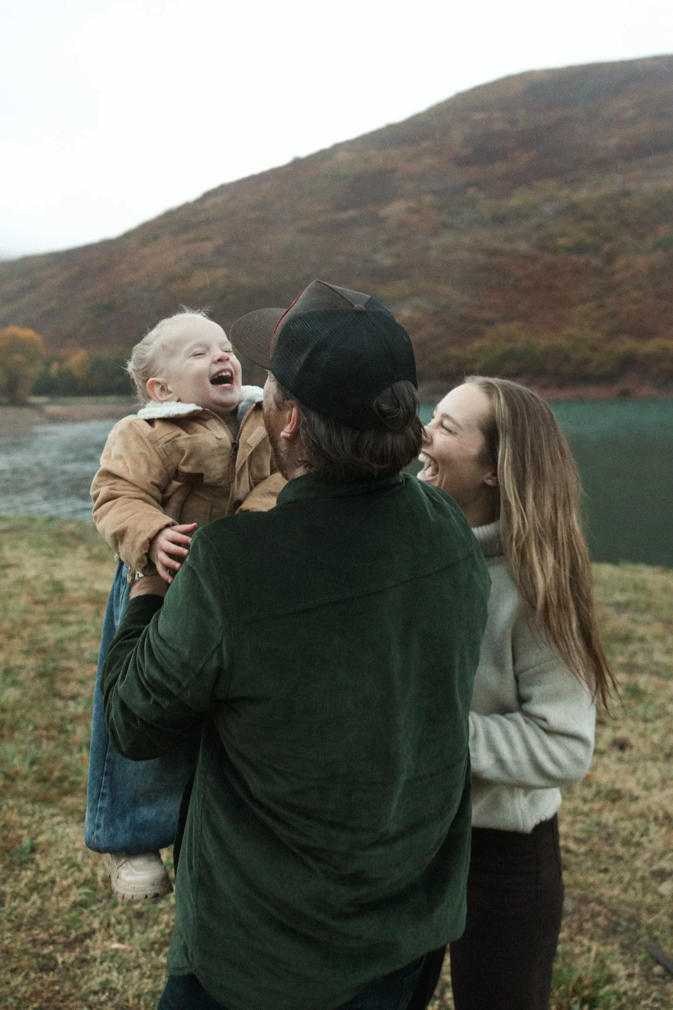 A family outdoors near a body of water with mountains in the background. A man is holding a young girl, both laughing and smiling, as a woman stands beside them, also smiling and enjoying the moment.