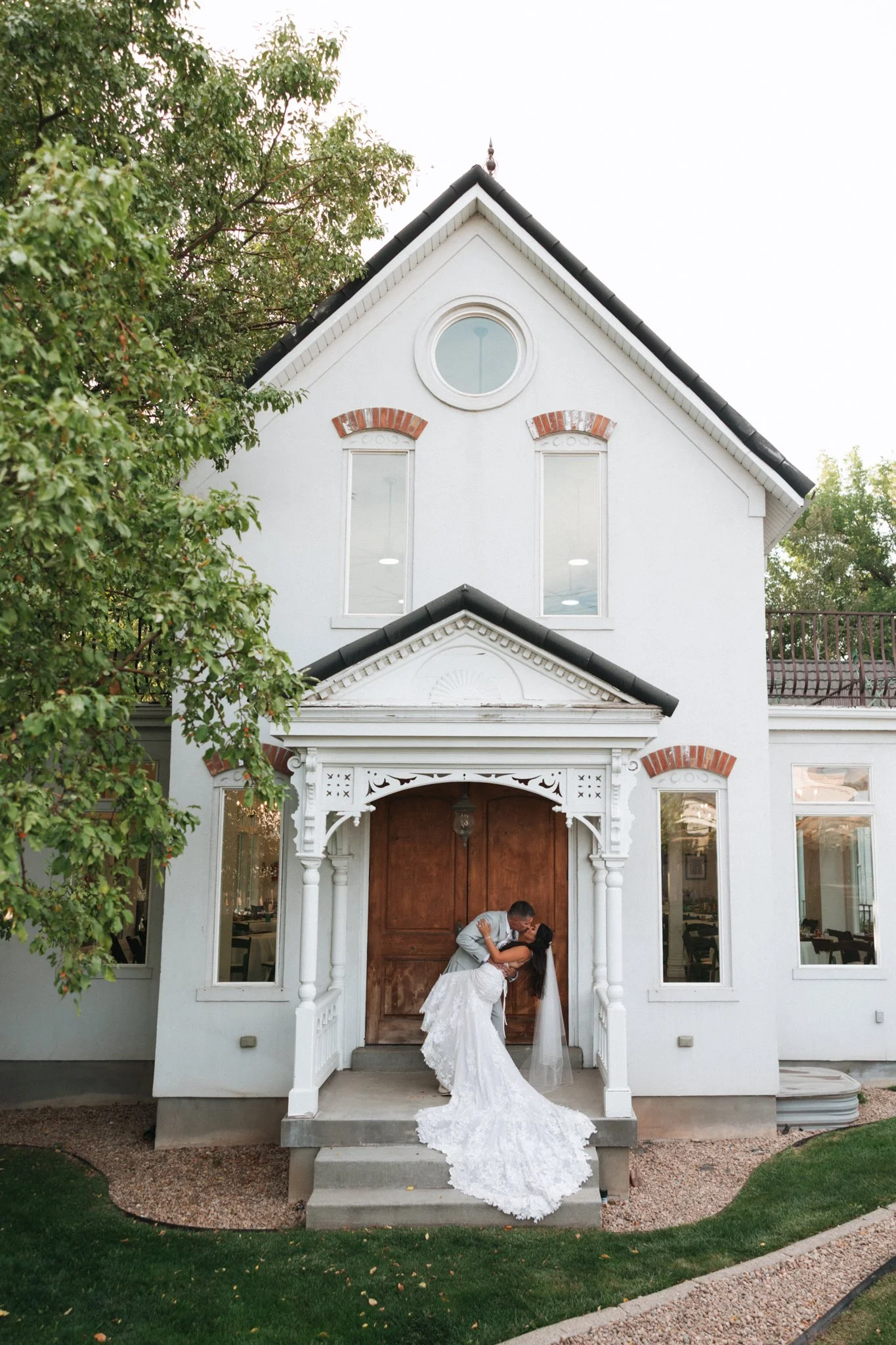 A bride and groom sharing a kiss in front of a white house with Victorian architecture, surrounded by greenery.