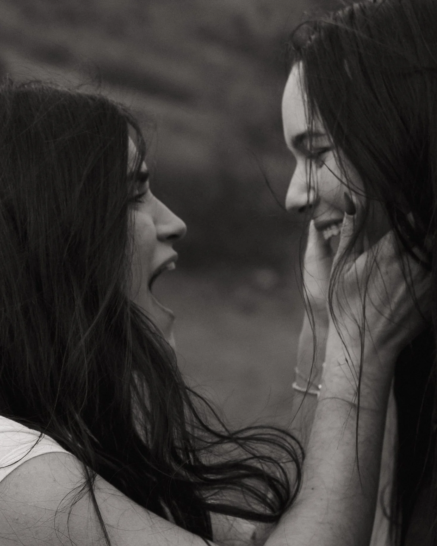 Two women with long dark hair are facing each other closely, smiling, in a black-and-white photo.