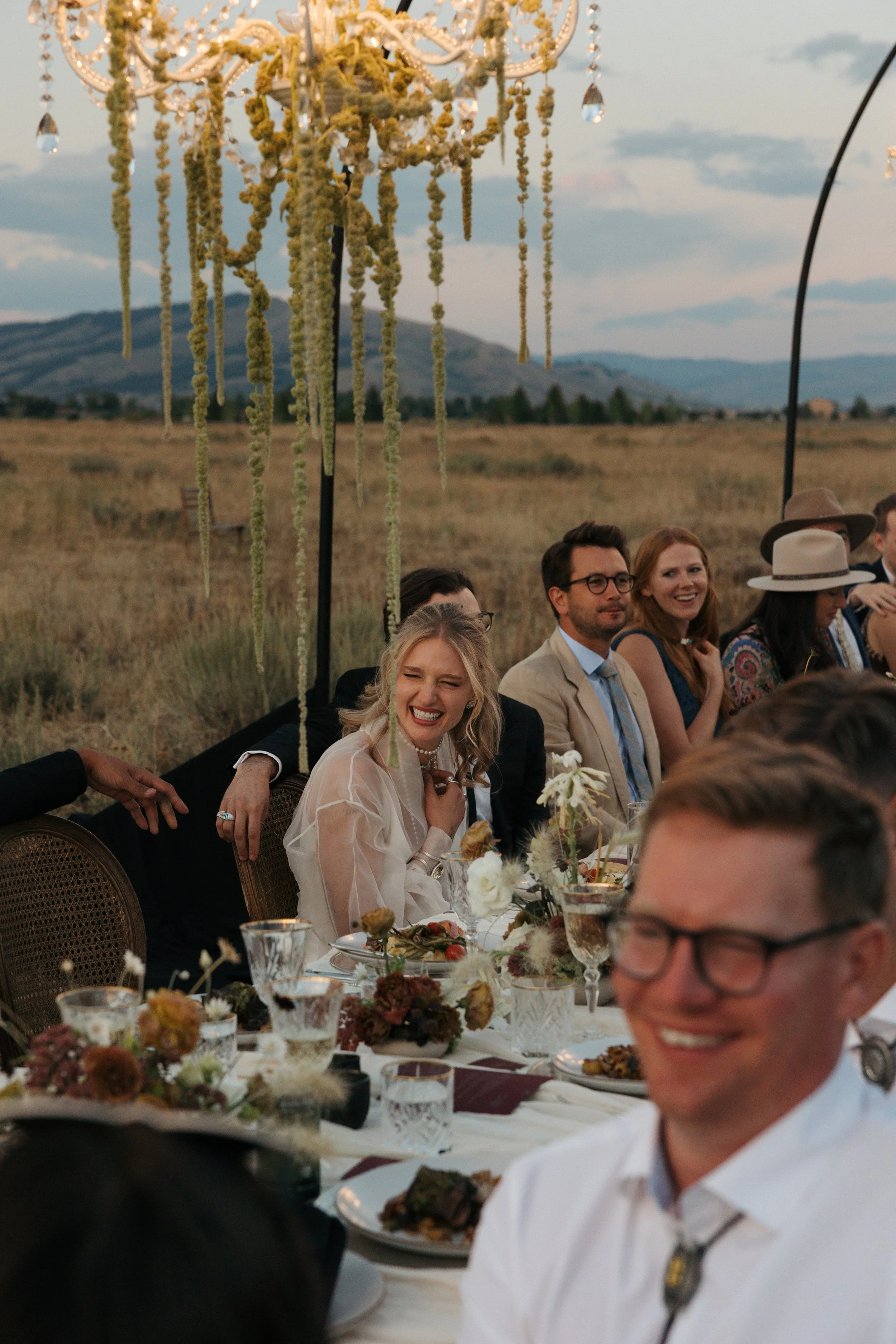 People seated at an outdoor wedding reception table with a scenic mountain view in the background, featuring a chandelier hanging overhead.