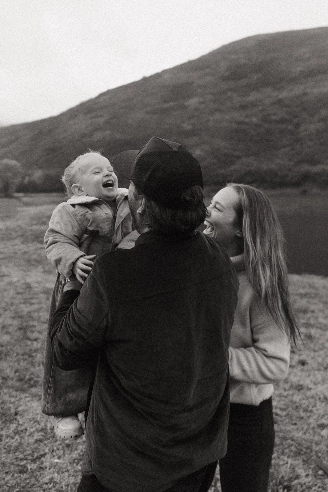 A family outdoors in a grassy field, with hills in the background, laughing and enjoying a happy moment. The child is being held up by a person with a baseball cap, and a woman with long hair is standing next to them.