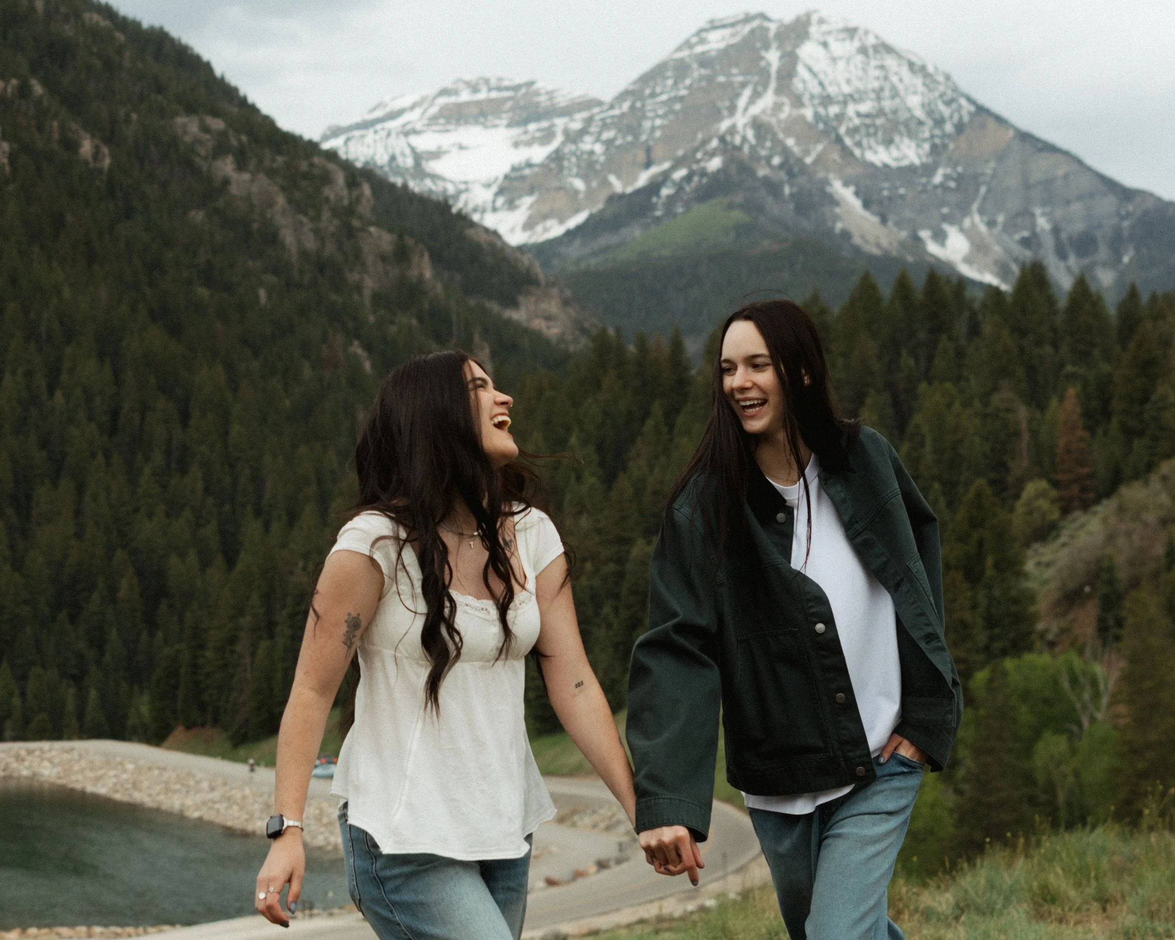 Two women walking hand in hand outdoors with snow-capped mountains and a forest in the background.