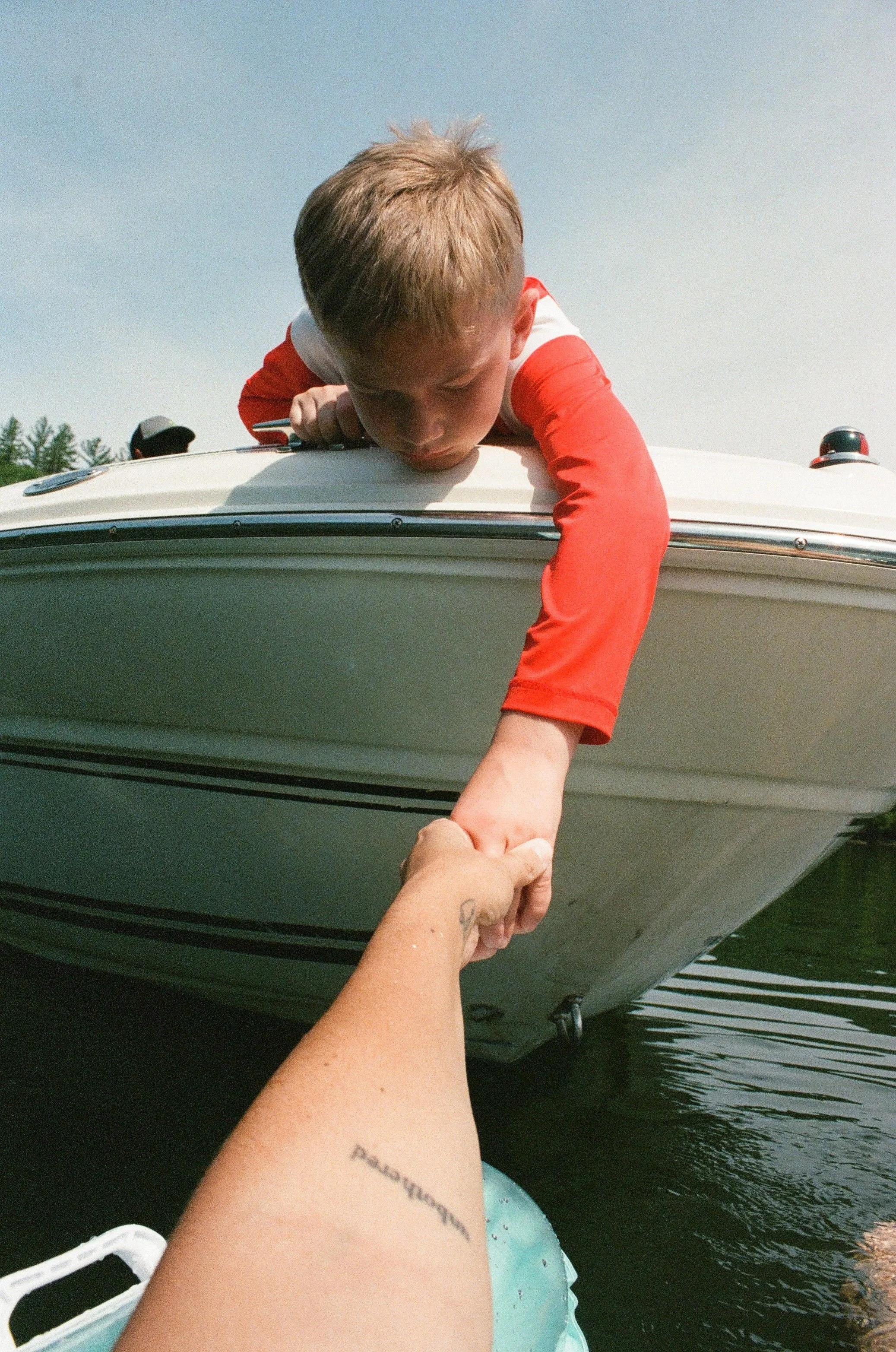 A person with tattoos is reaching out from a boat to hold the hand of a child who is leaning over the boat's edge. The child is wearing a red and white shirt and is looking down. The boat is on the water, with another person visible in the background.
