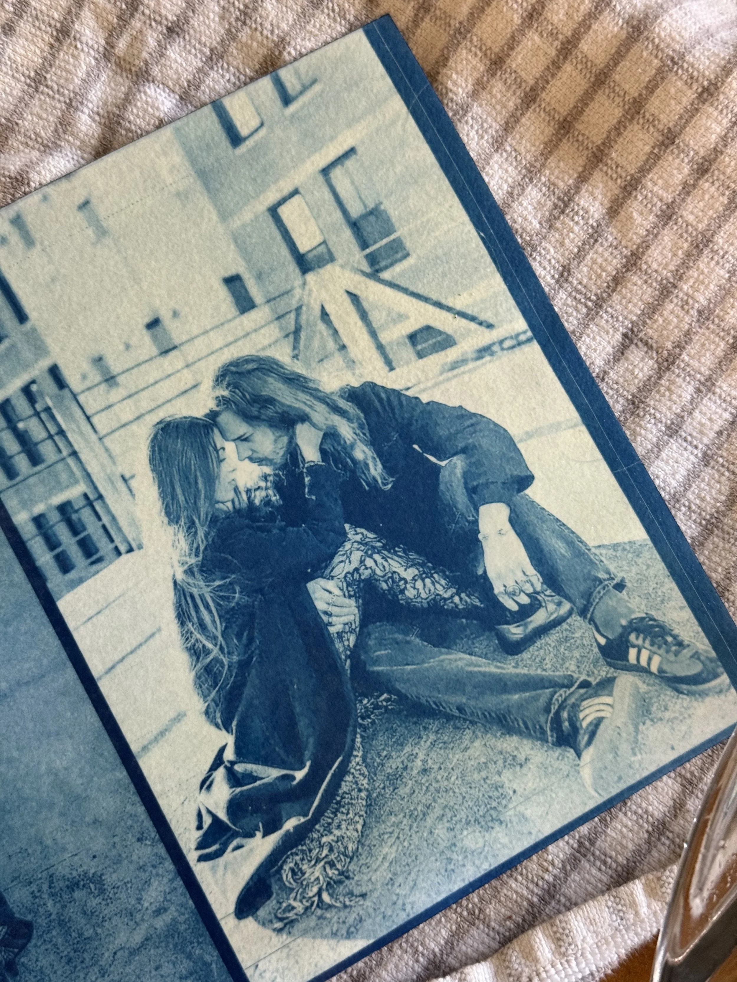 Black-and-white photo of a young couple sitting on the ground, leaning close to each other, with city buildings in the background.