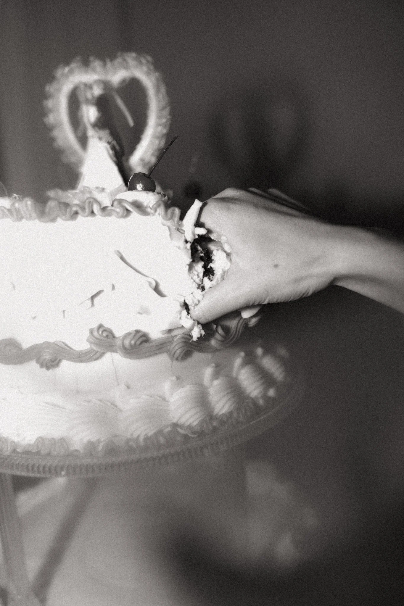A birthday cake with white frosting, decorated with a cherry, a heart-shaped cookie, and a lit candle, on a clear plate.