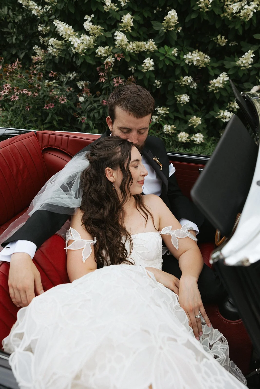 A bride and groom are sitting close together in a vintage car, sleeping or resting with their eyes closed. The bride is wearing a white dress with bow details on her shoulders and a veil, and has long dark wavy hair. The groom, dressed in a black tuxedo, has his head gently resting on the bride's head. They are surrounded by greenery and white flowers outside the car.