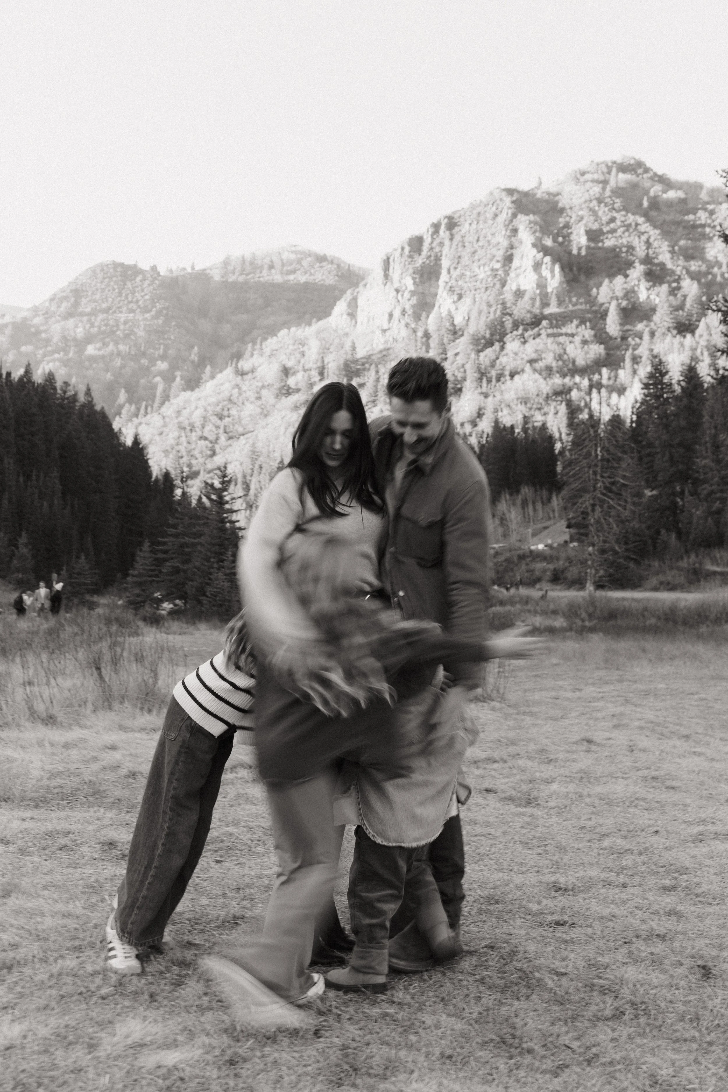 A black and white photo of a group of people dancing and celebrating outdoors with mountainous terrain in the background.