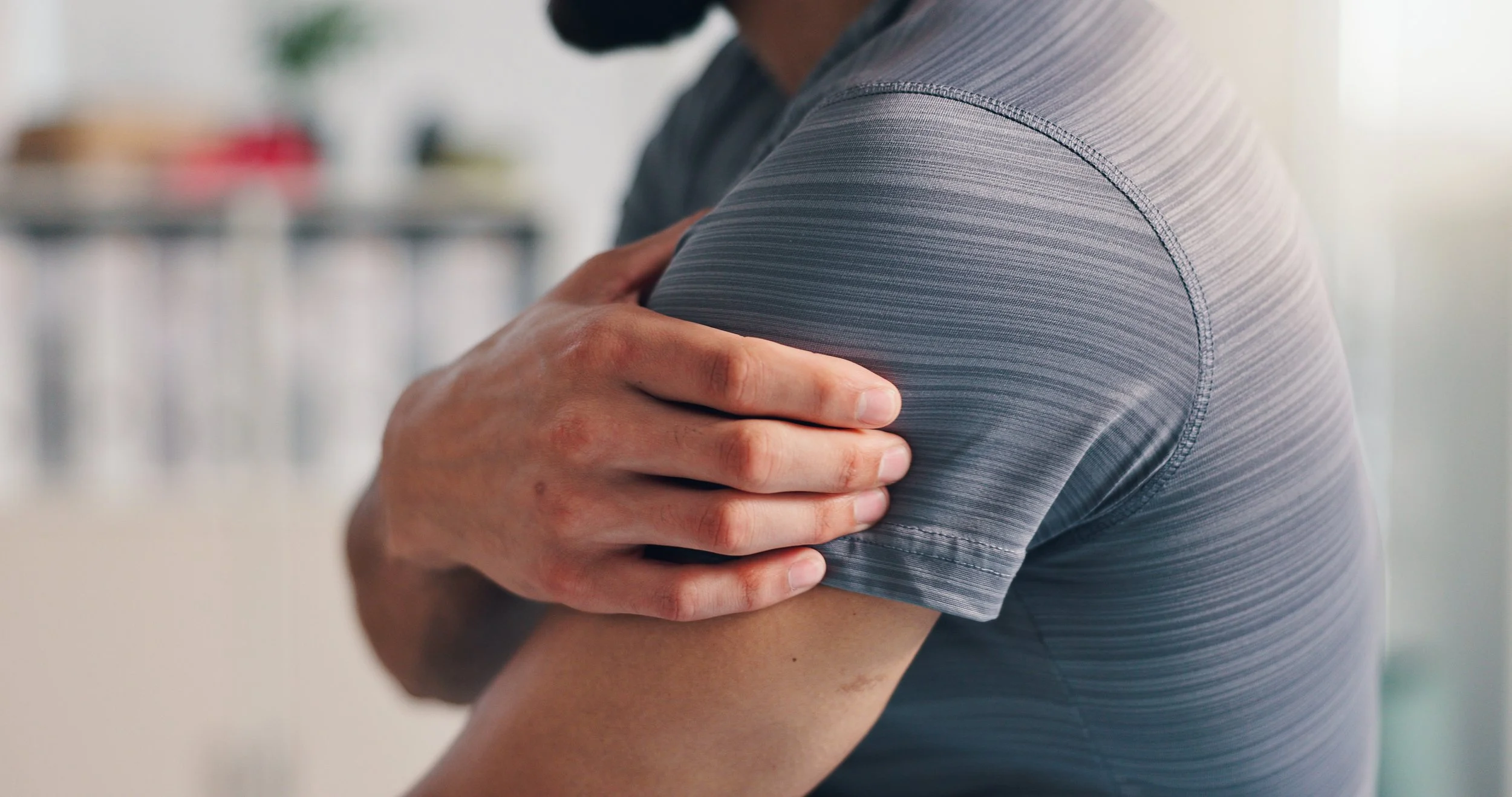 A person wearing a gray athletic shirt holding their shoulder with one hand, possibly indicating discomfort or pain.